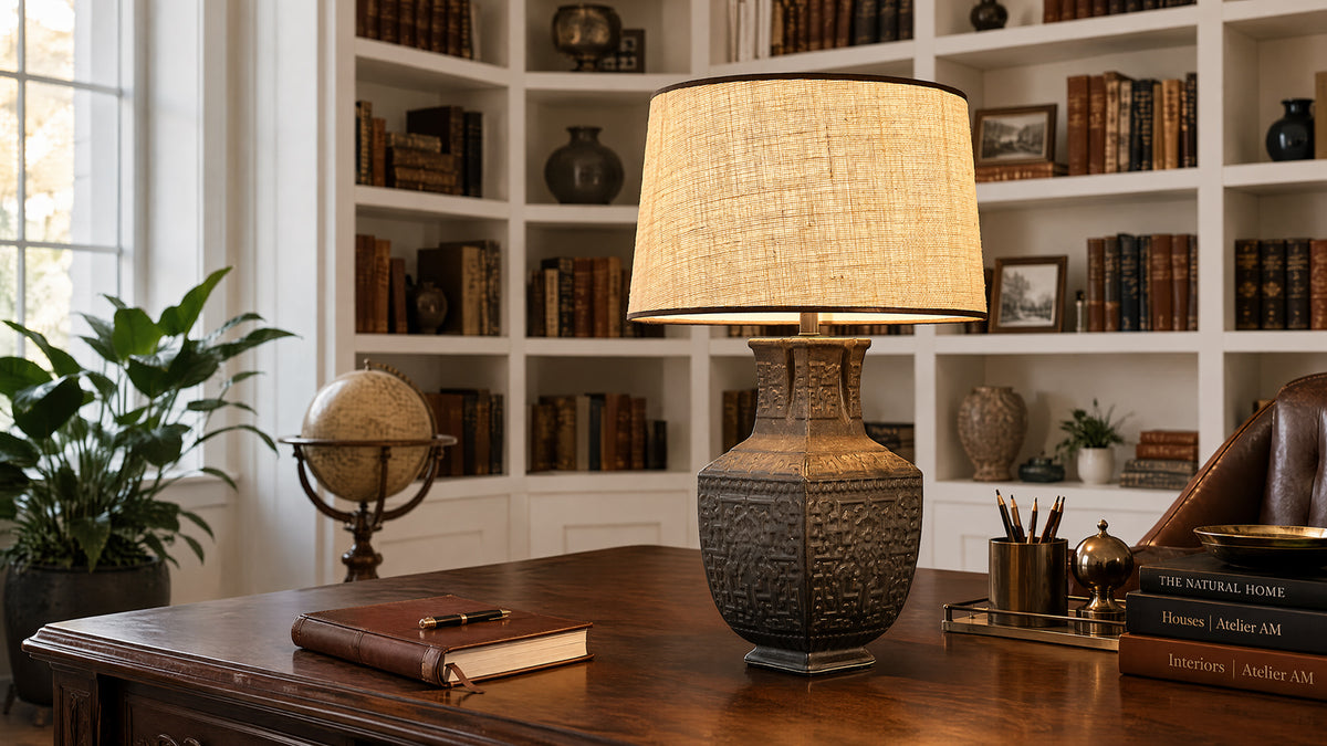 Artisanal carved ceramic vessel table lamp glowing softly on a rich wooden desk in a classic home office with filled bookshelves.