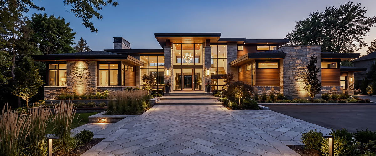 A panoramic evening view of a mid-century modern villa wrapped in lush greenery, with warm, layered interior lighting glowing through large glass walls against a dramatic dusk sky.