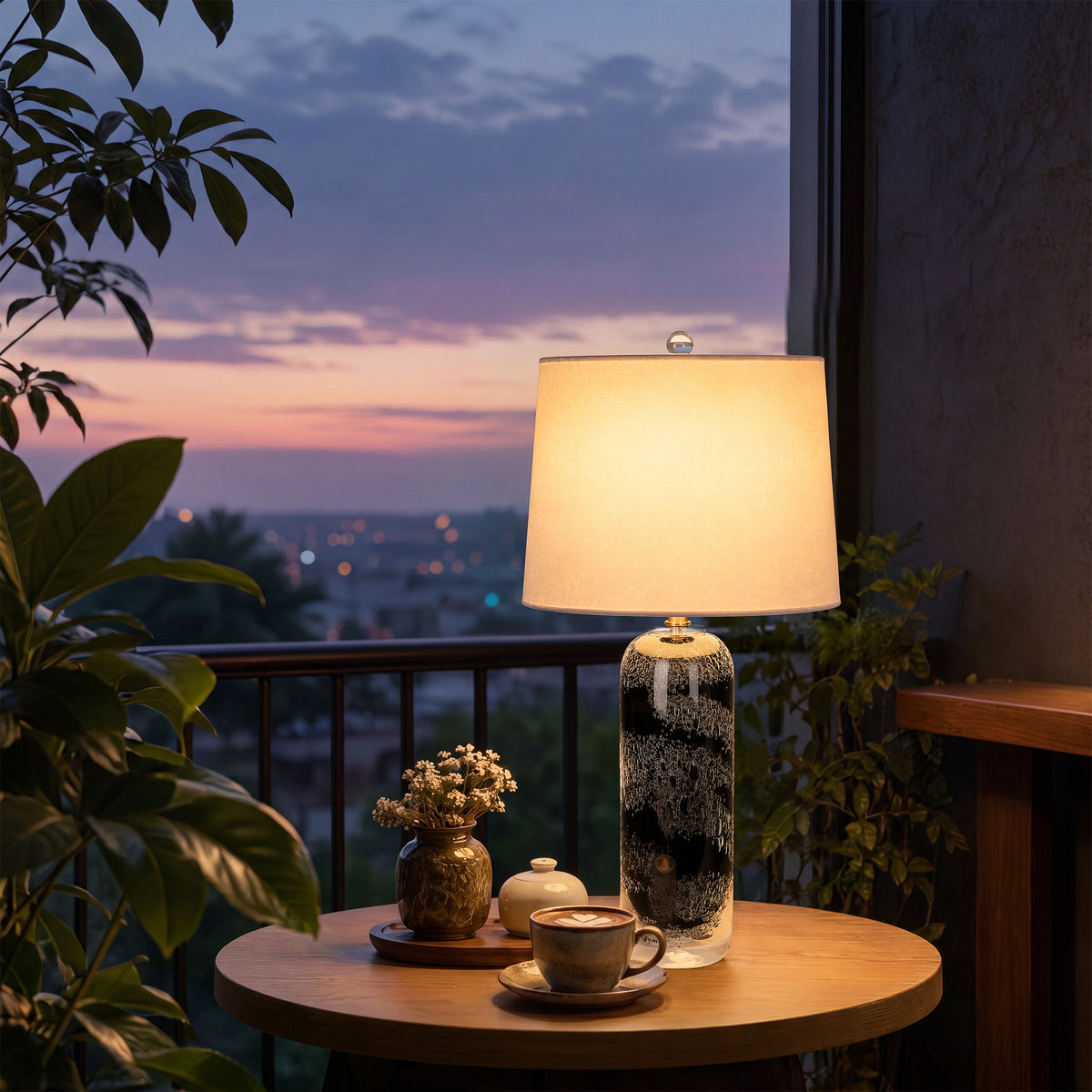 Art glass table lamp illuminated on a round balcony coffee table against a dusk sky and city view.