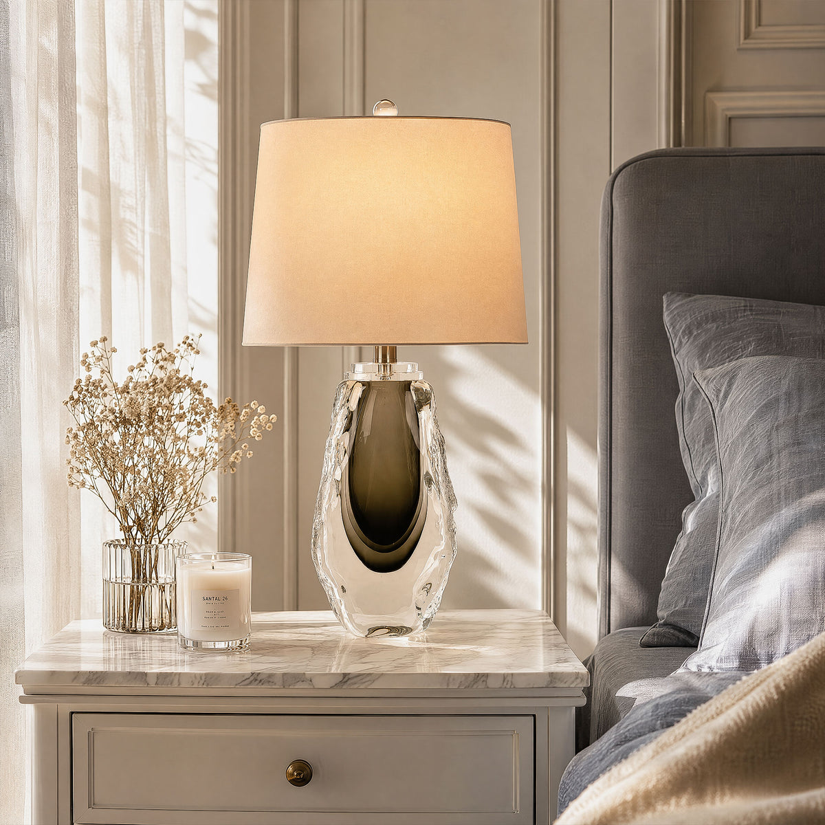 Art glass table lamp with a warm glowing shade on a marble bedside table in a sunlit bedroom setting.