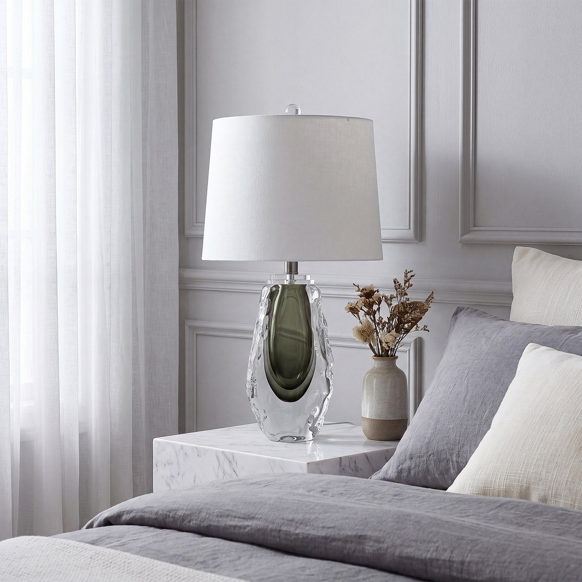 Unlit sculptural art glass table lamp resting on a white marble bedside table next to a bed with grey linens.