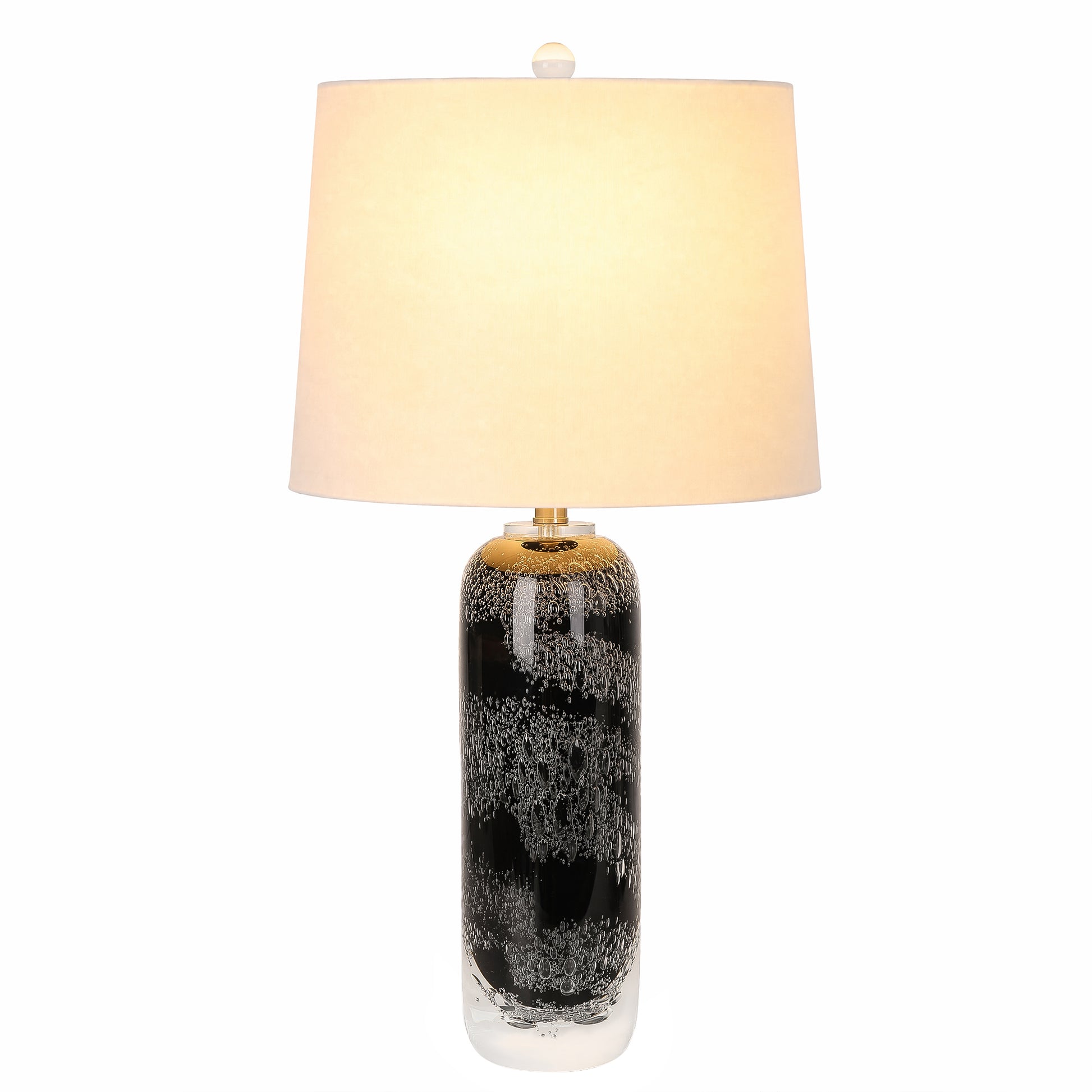 Art glass table lamp with black seeded glass body glowing with warm light beneath a white fabric shade on a plain background.