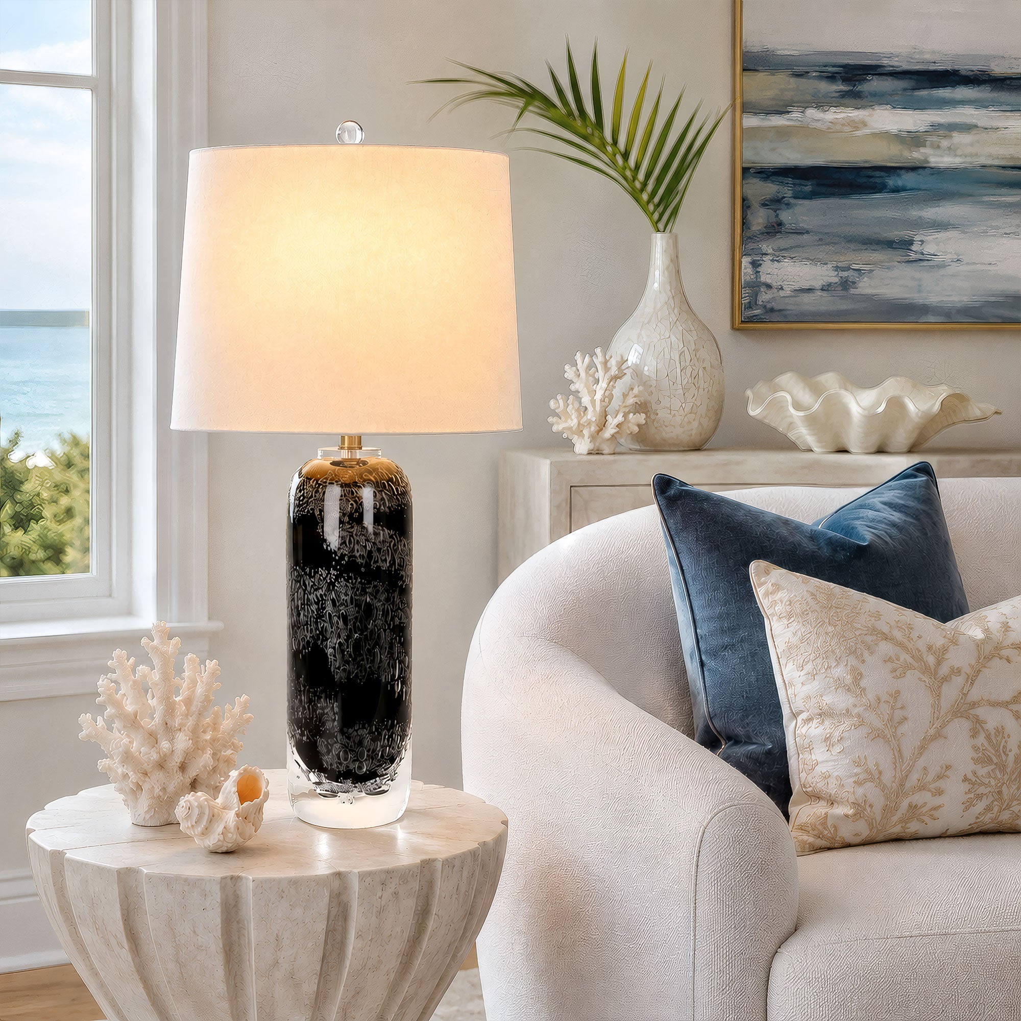 Art glass table lamp casting warm light on a stone side table beside a sofa in a coastal styled living room.