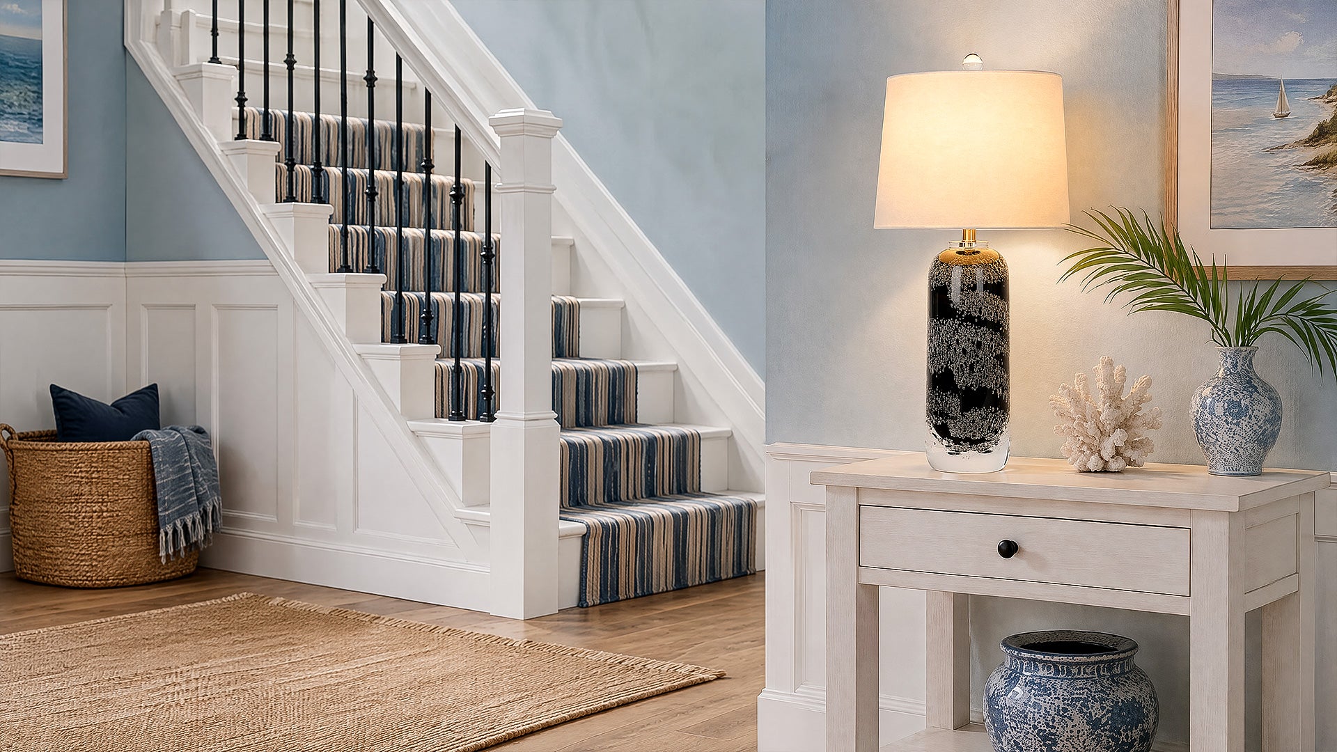 Warmly lit art glass table lamp placed on a white console table in an elegant entryway foyer next to a staircase.