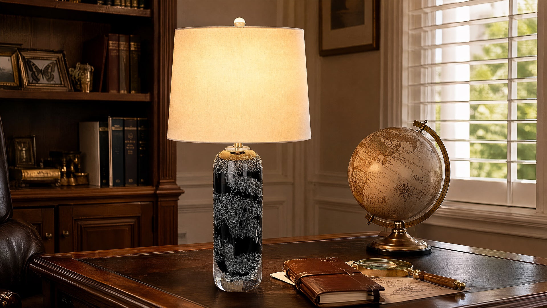 Illuminated seeded art glass table lamp standing on a luxury dark leather desk next to a classic globe in a home office.