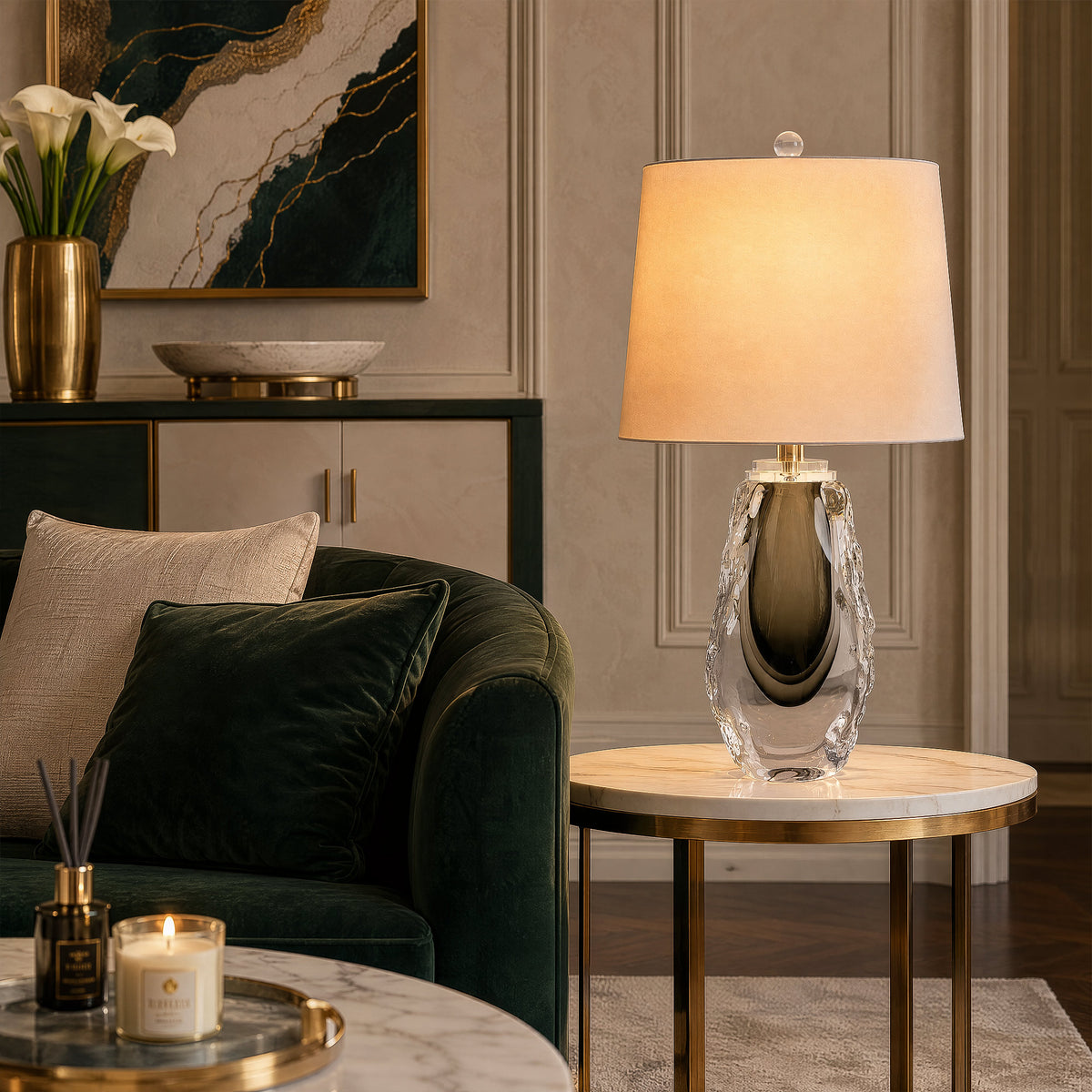 Lit art glass table lamp sitting on a round marble end table near a dark green sofa in a modern living room.