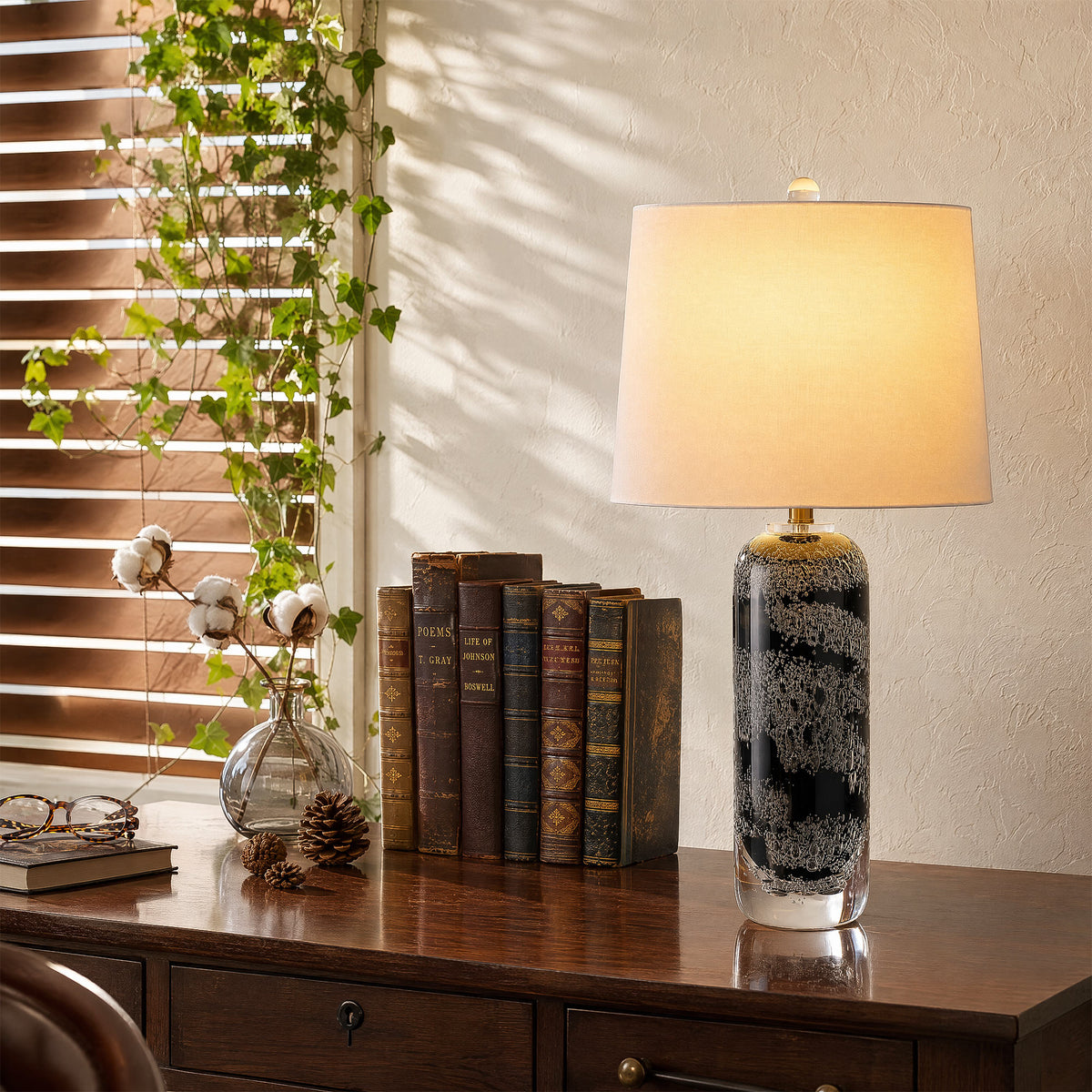 Warm illuminated art glass table lamp with bubble texturing placed next to vintage books on a wooden desk near a window.
