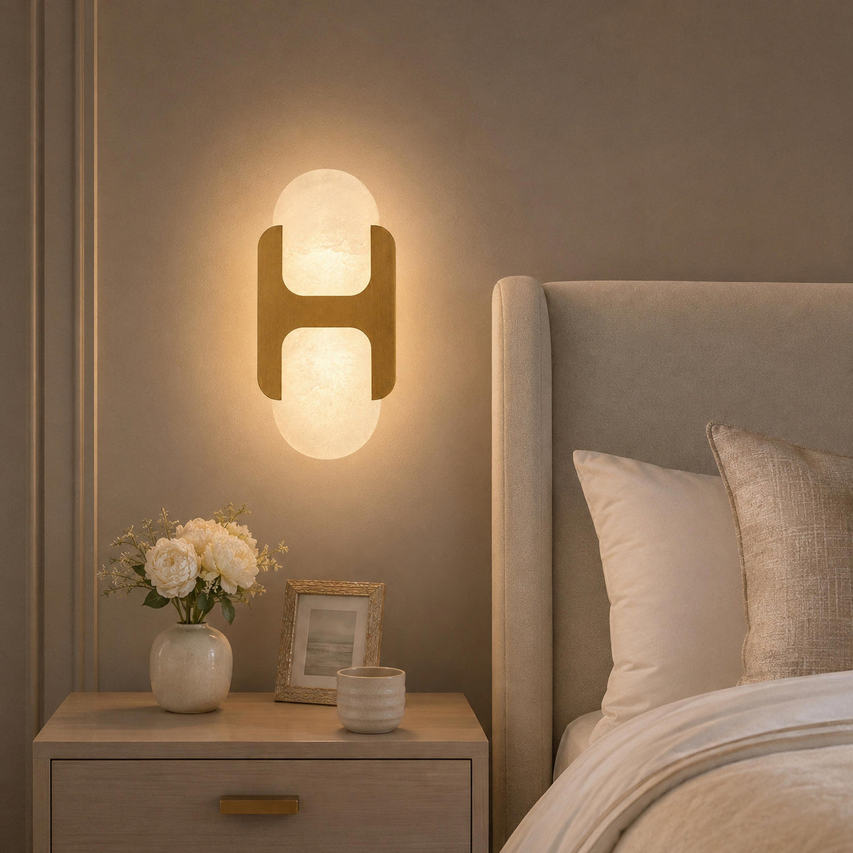 Warm illuminated H-shaped alabaster wall sconce mounted beside a upholstered bedhead above a nightstand in a cozy bedroom