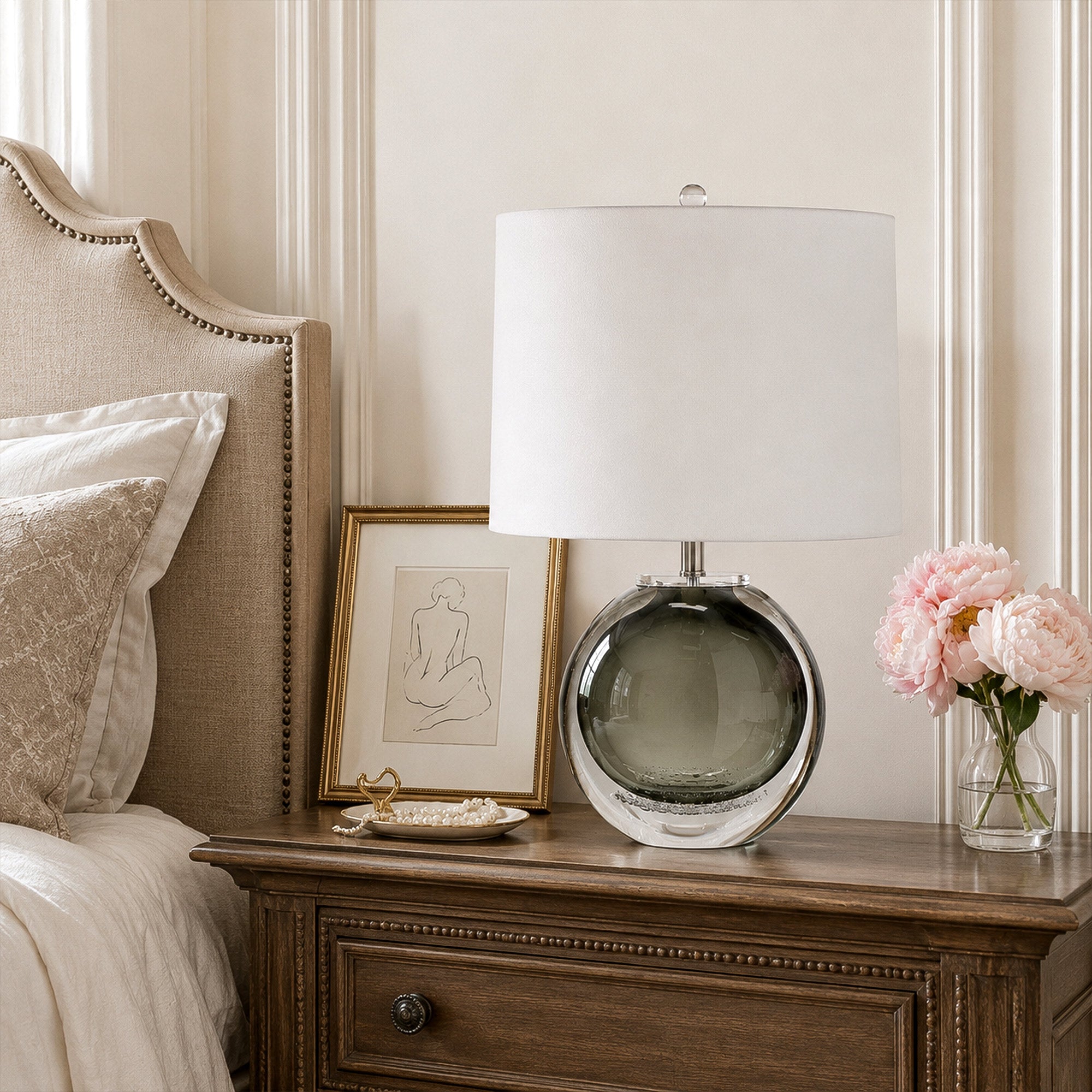 Smoke gray art glass table lamp with white drum shade placed on a dark wood nightstand in an elegant bedroom