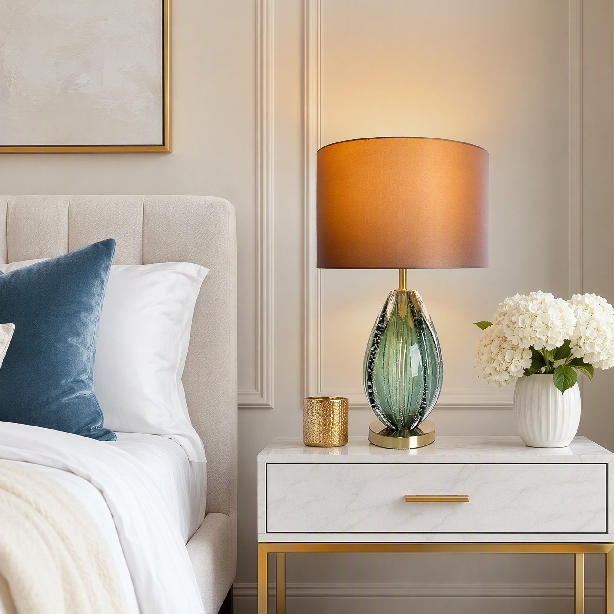 Illuminated teal art glass table lamp on white marble top nightstand beside upholstered bed with blue pillow