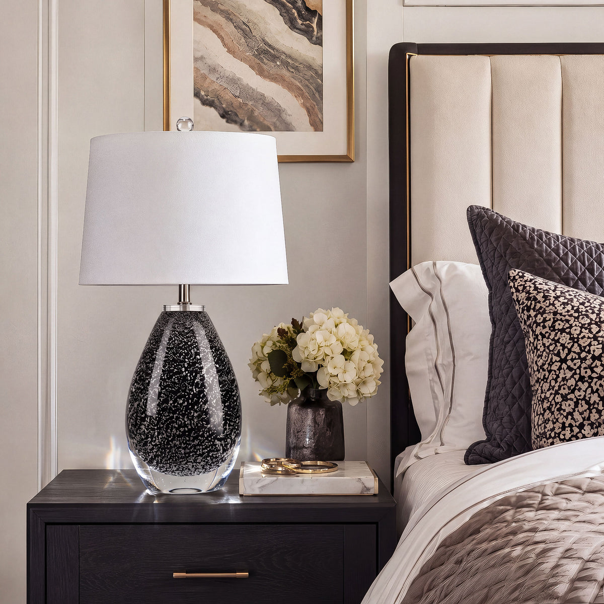 Teardrop black art glass table lamp on black nightstand beside upholstered bed in modern bedroom.