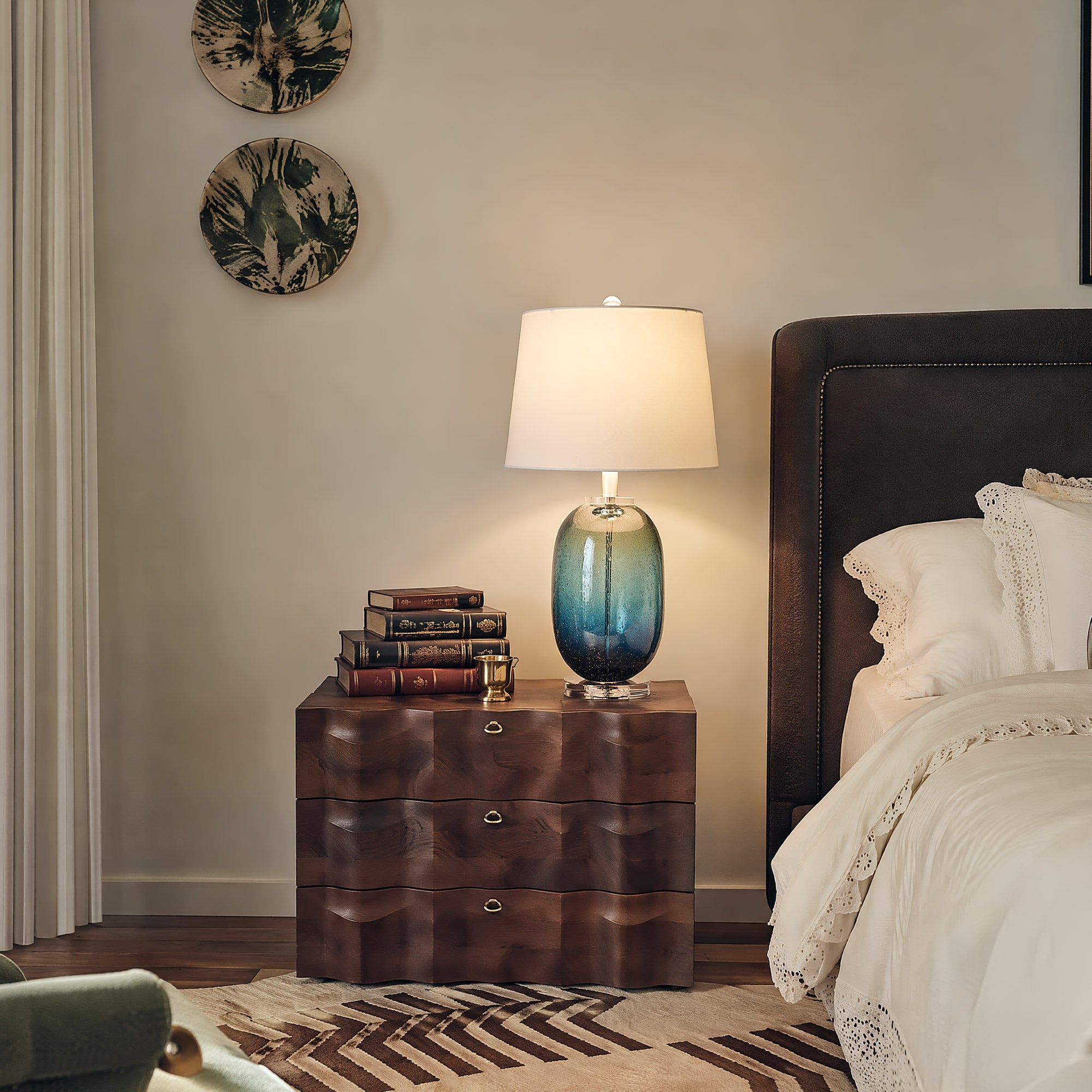 A sleek barrel shaped blue ink bubble glass bedside lamp illuminating a wave pattern dark wood nightstand next to a luxury tufted bed