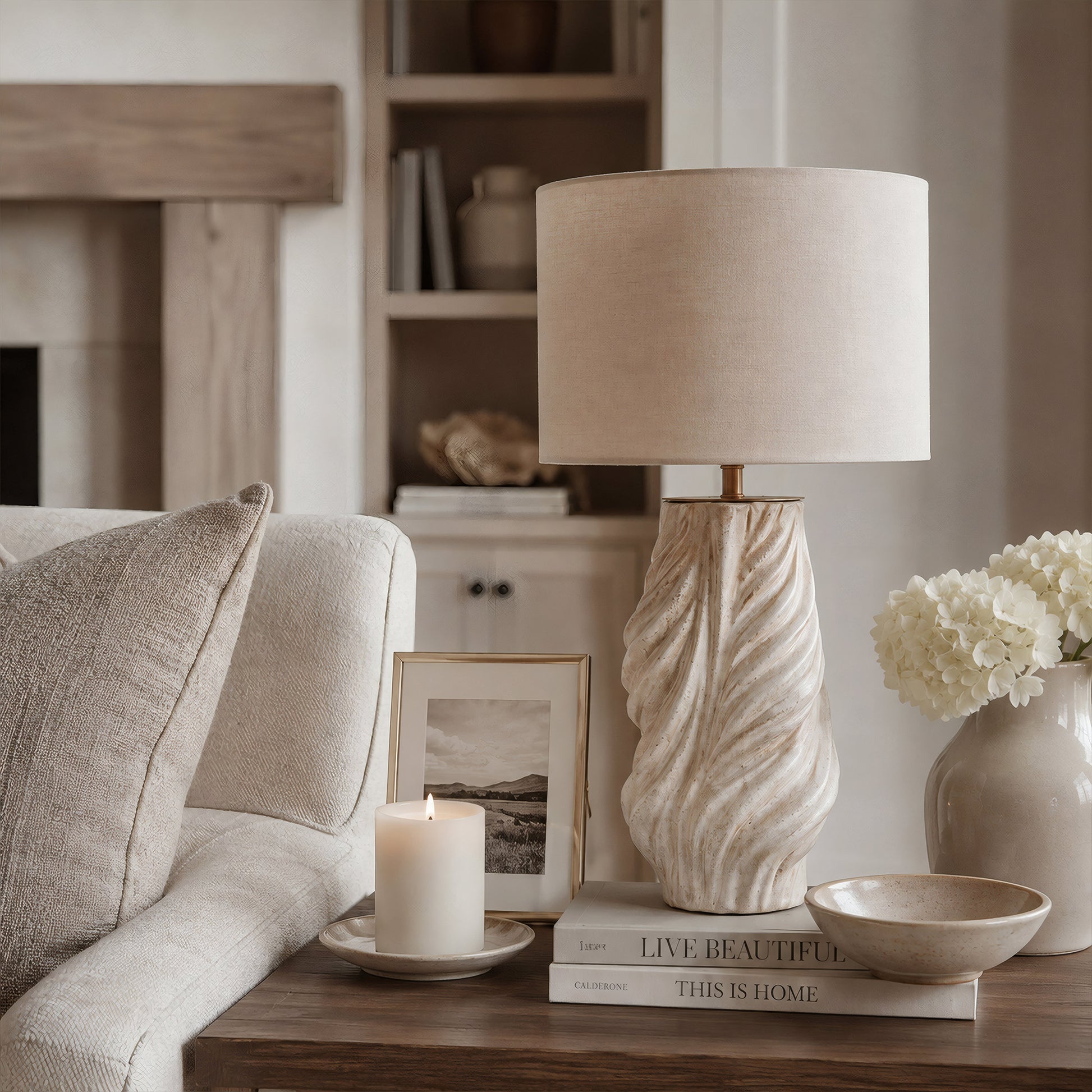 A white grey fluted ceramic table lamp resting on a wooden side table next to a cream fabric sofa and fresh white flowers