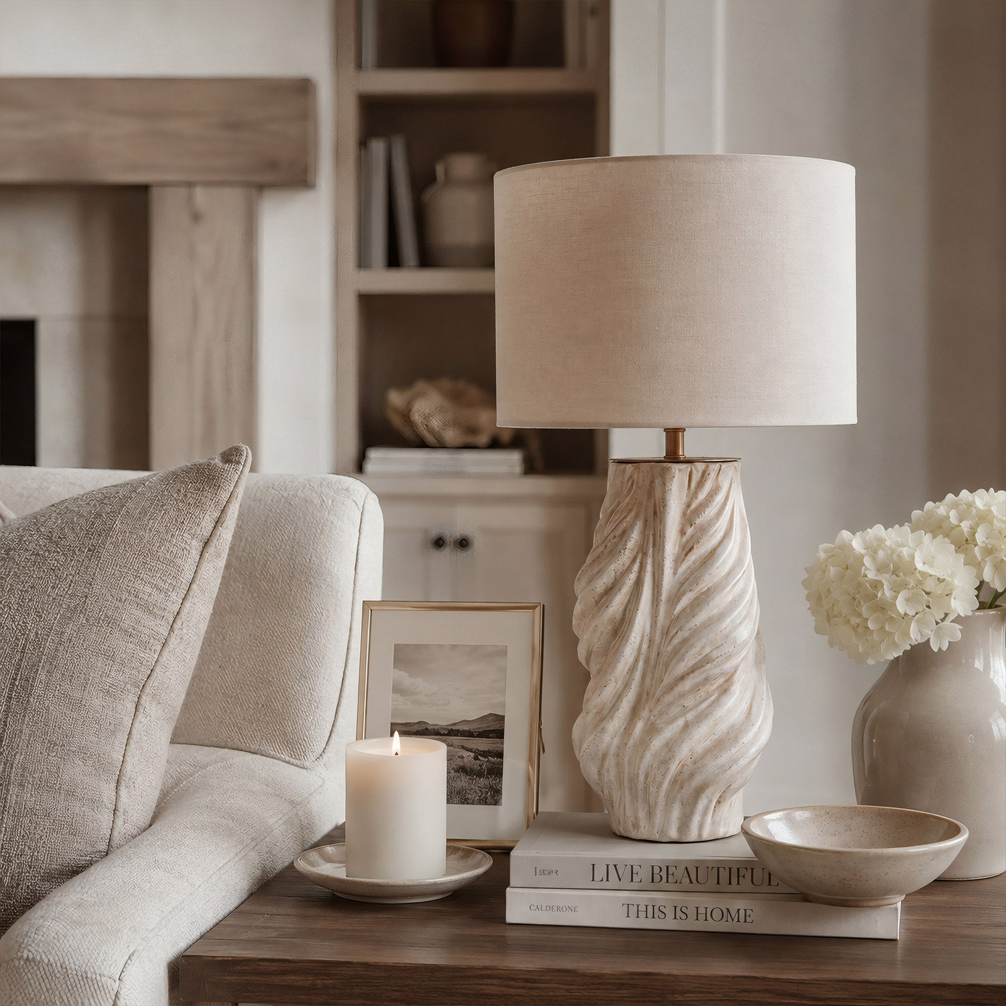 A white grey fluted ceramic table lamp resting on a wooden side table next to a cream fabric sofa and fresh white flowers