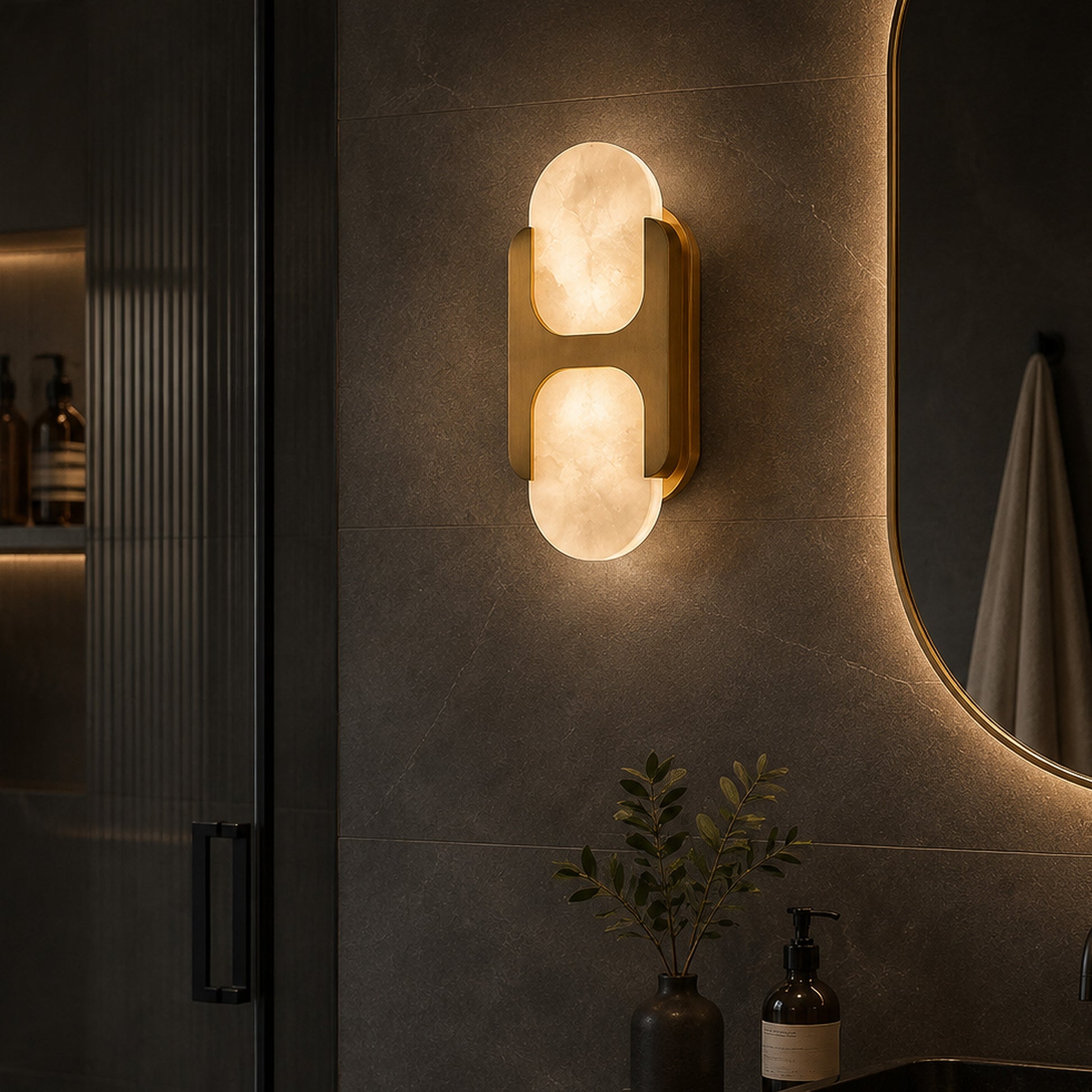 Warmly lit brass and Spanish alabaster wall sconce mounted on dark tiled wall beside a mirror in a modern moody bathroom