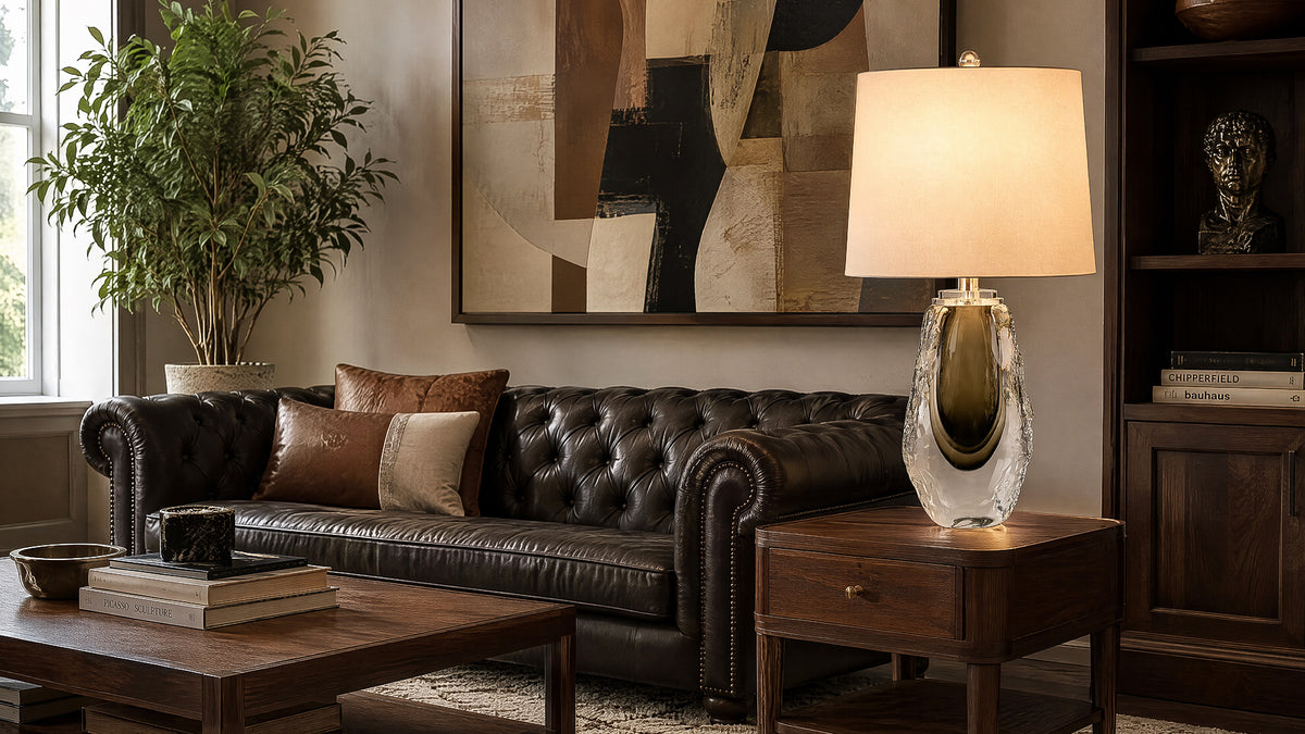 Art glass table lamp lit on a dark wood end table next to a leather sofa in a dark interior.