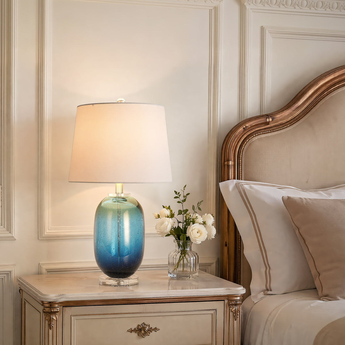 Warmly lit tall barrel blue ink art glass bedside lamp resting on an elegant cream provincial nightstand with white roses in a French style bedroom