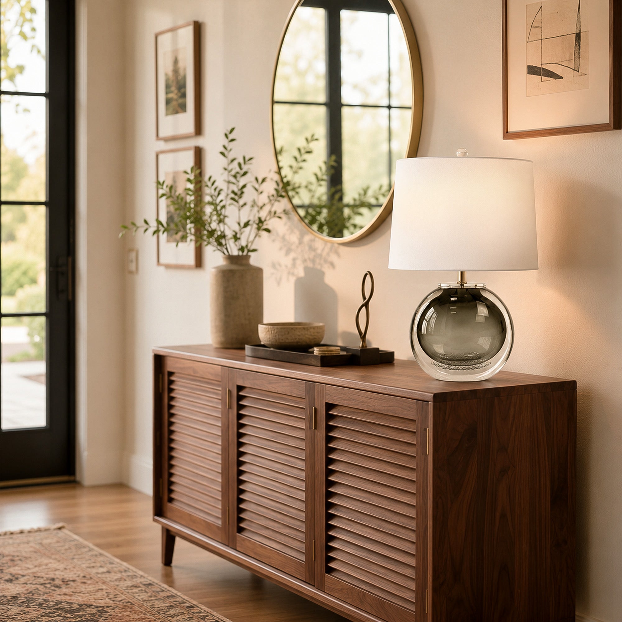 Illuminated art glass table lamp on a dark wood console table in a bright entryway with round mirror