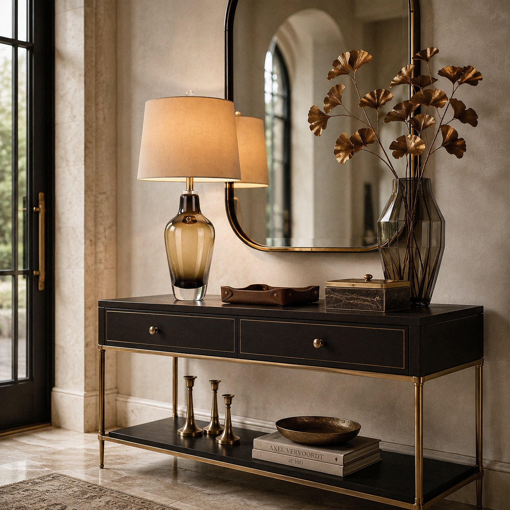 Illuminated art glass table lamp on black console table with gold legs and large arched mirror in entryway