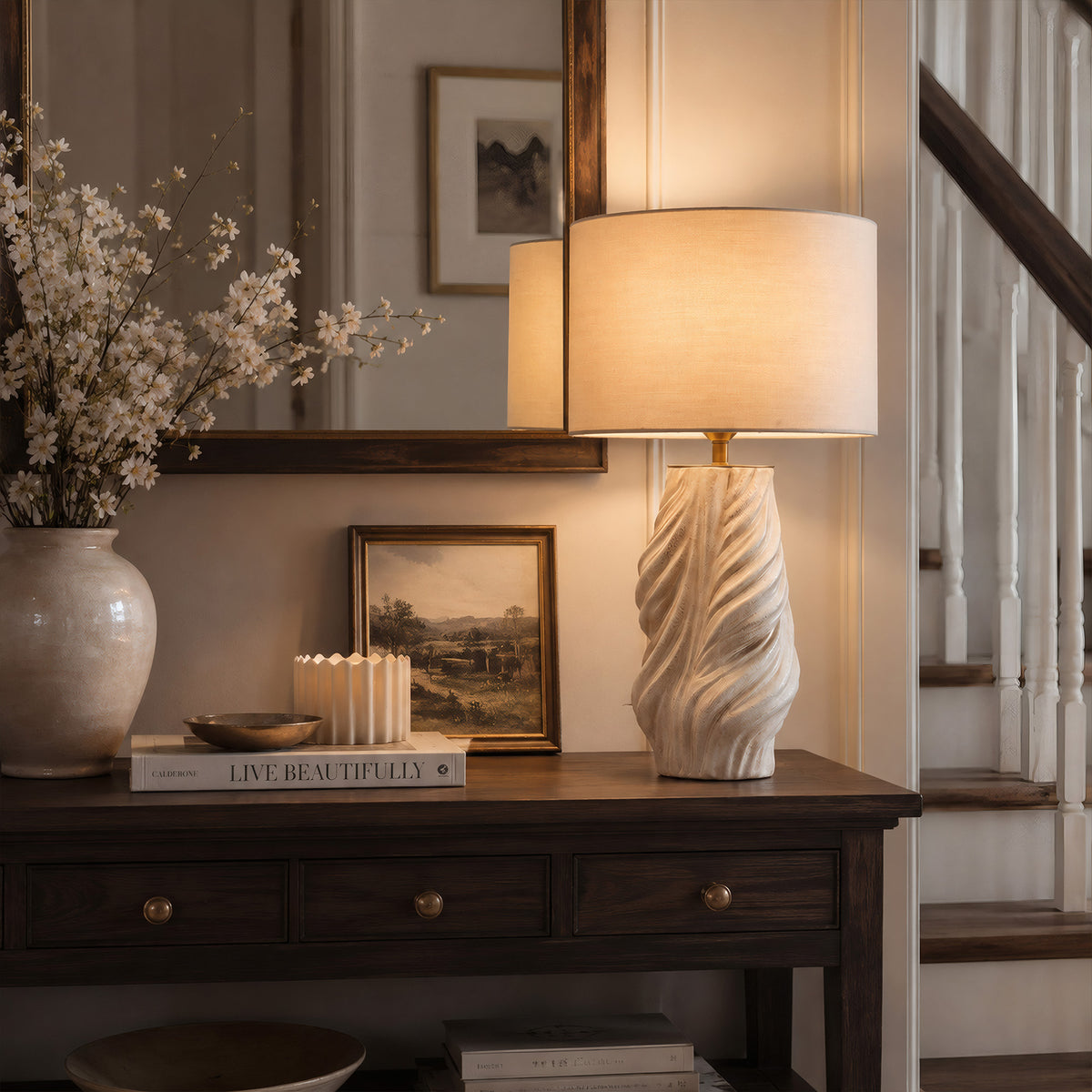 An elegant entryway console table featuring a lit white and grey twisted ceramic lamp next to a vase with white flowers and a round mirror