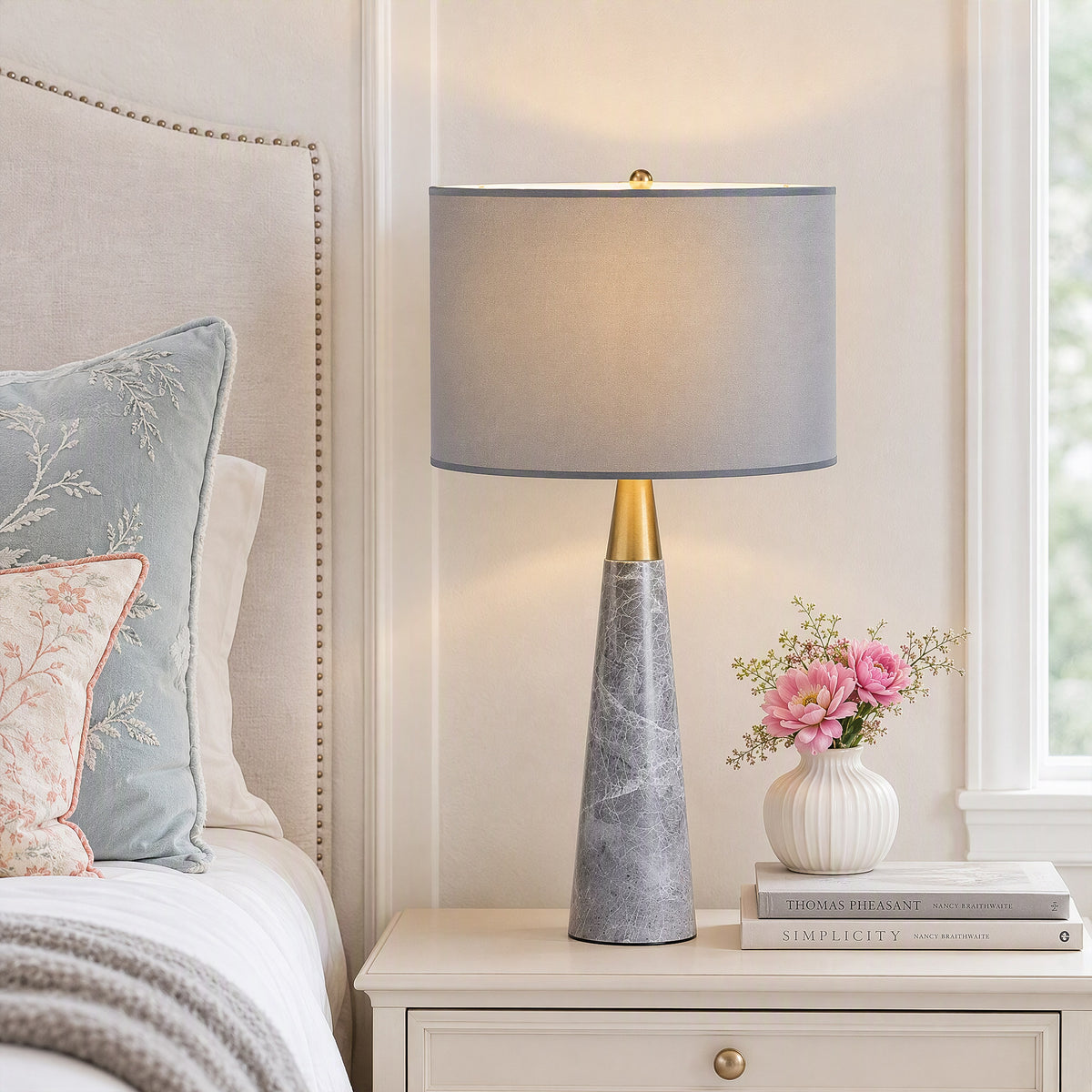 Illuminated grey marble cone table lamp on white nightstand with pink flowers in vase