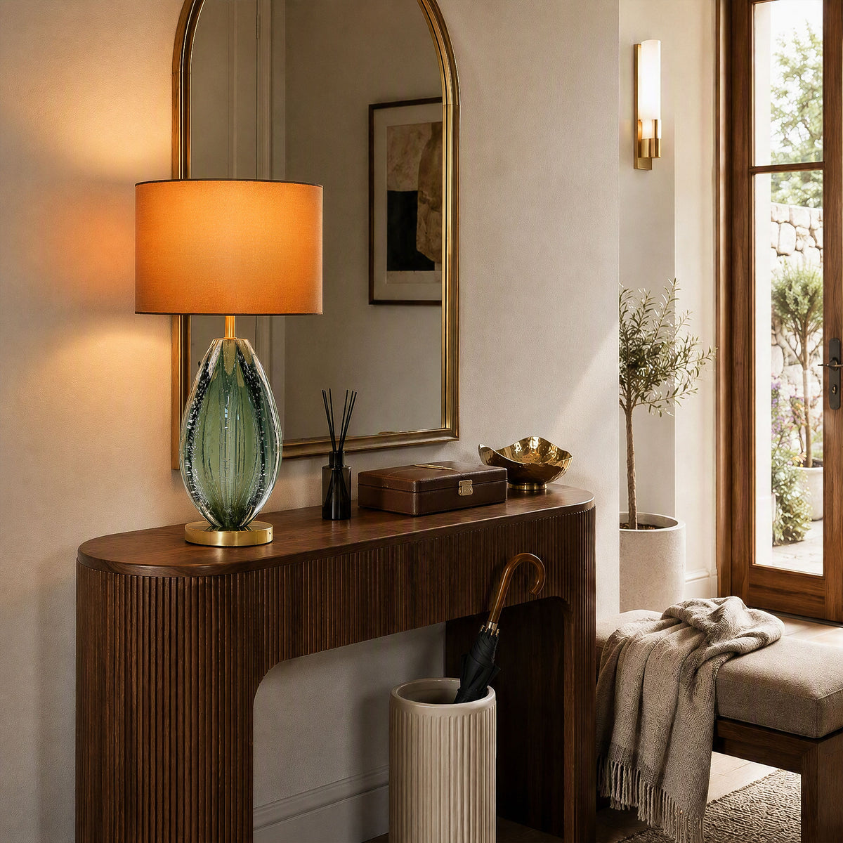 Illuminated teal seeded glass table lamp on wooden entryway console table in front of arched brass mirror