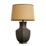 Handcrafted vintage vessel ceramic table lamp with carved relief patterns and burlap shade on white background with light off