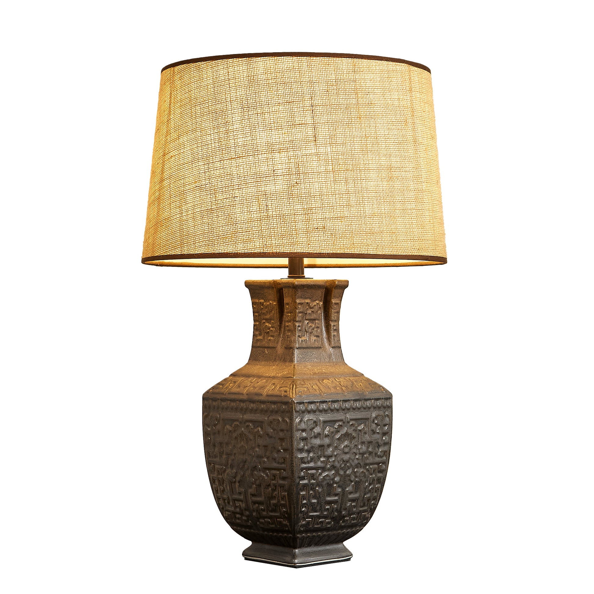 Illuminated handcrafted vintage vessel ceramic table lamp with warm glow through burlap shade on white background