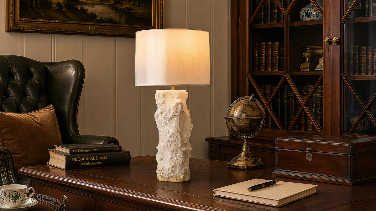 Illuminated organic ceramic vessel table lamp sitting on antique wooden desk beside globe in traditional home study