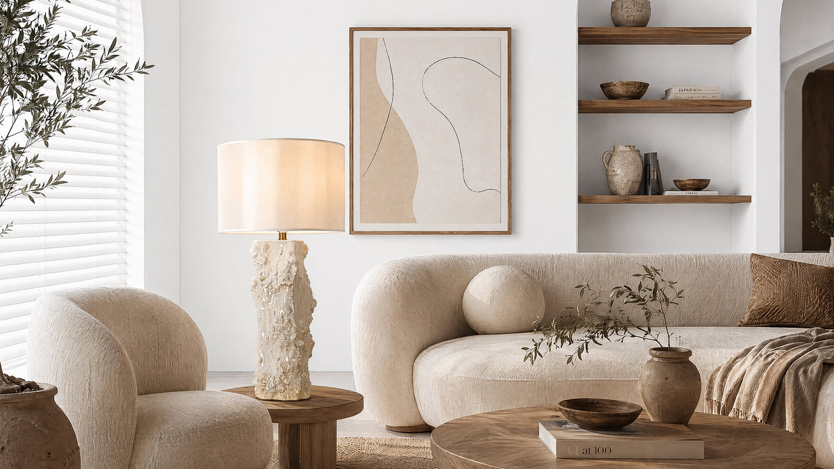 Warmly lit handmade ceramic vessel table lamp on wooden stool beside boucle armchair in bright minimalist living room