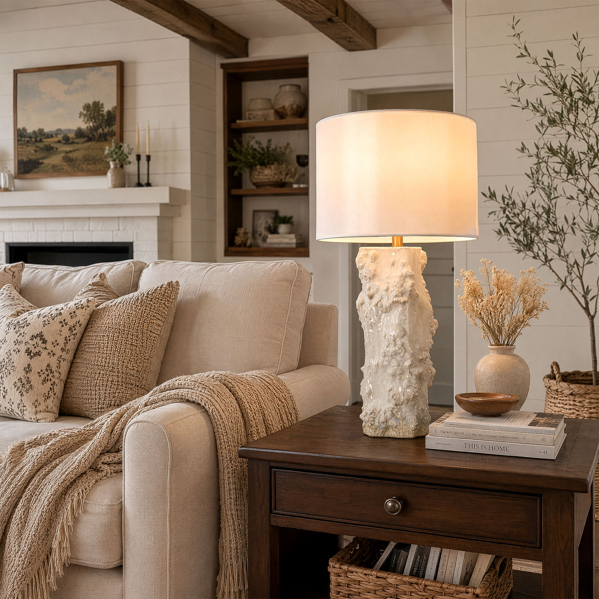 Illuminated handmade ceramic vessel table lamp sitting on dark wood end table next to beige sofa in living room