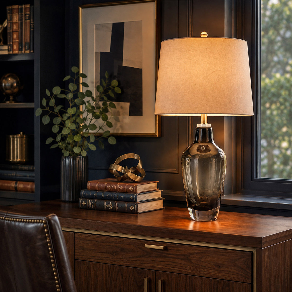Illuminated art glass table lamp on dark wood desk by window in sophisticated home office study