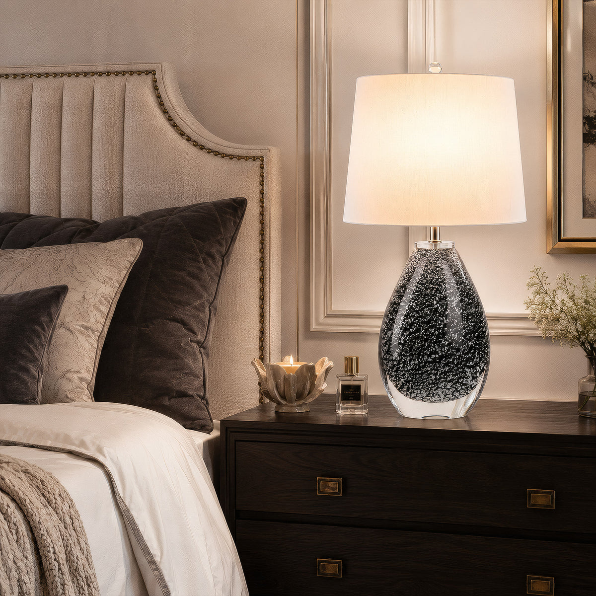 Illuminated teardrop glass table lamp casting soft light on wooden nightstand in warm bedroom interior.