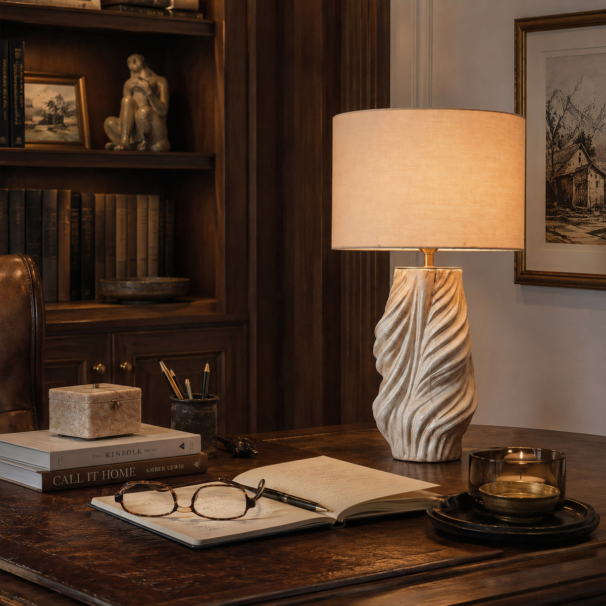 An illuminated sculptural white and grey twisted ceramic lamp providing warm task lighting on an executive wooden desk in a home office