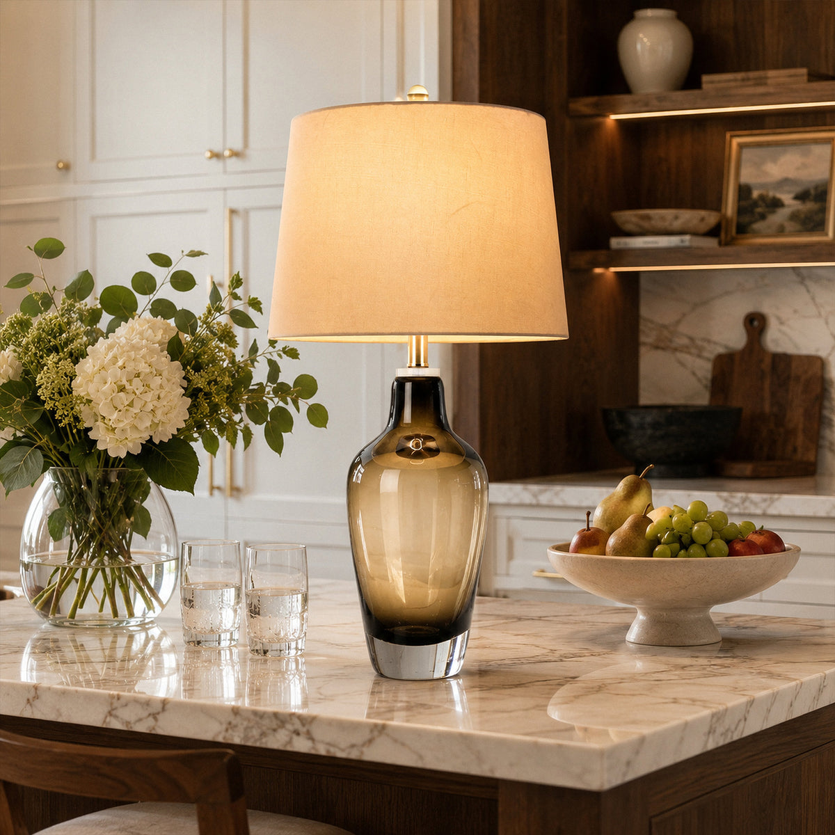 Illuminated art glass table lamp on marble kitchen island next to vase of flowers and fruit bowl