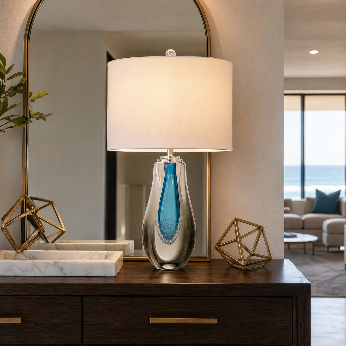 Art glass table lamp lit on a dark wood console table in a living room entryway with arch mirror