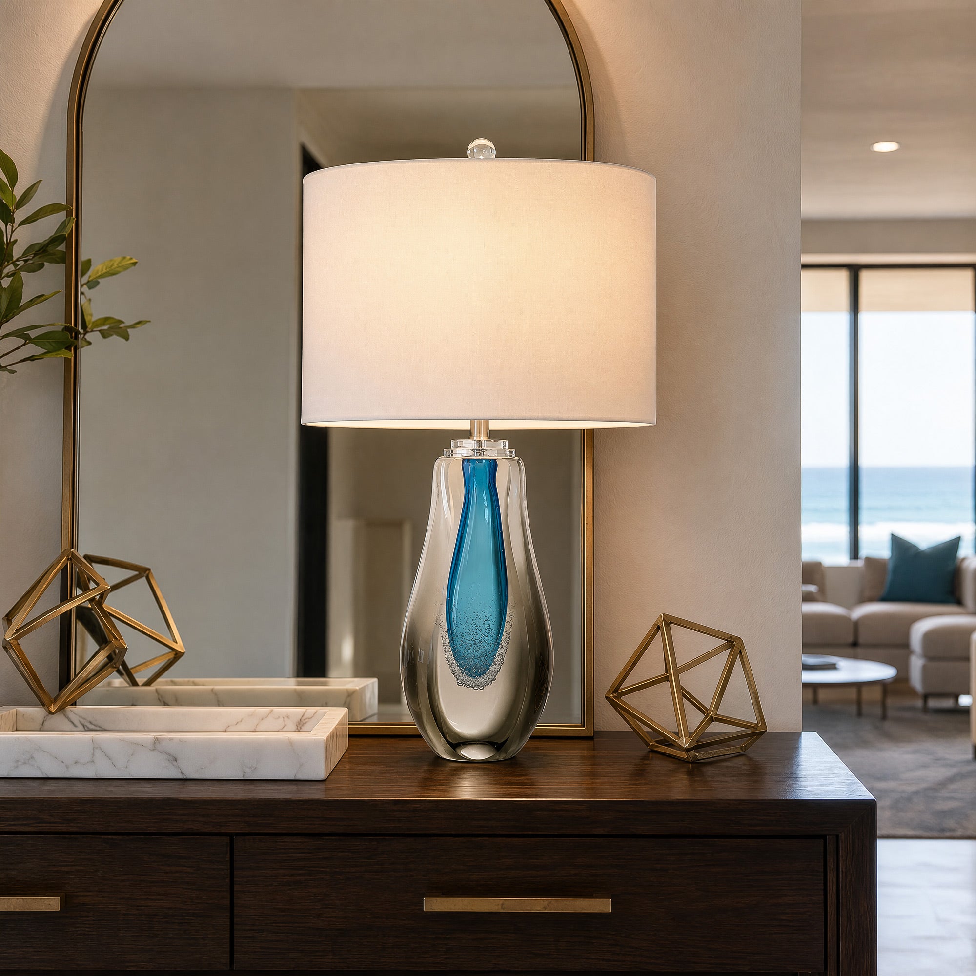Art glass table lamp lit on a dark wood console table in a living room entryway with arch mirror