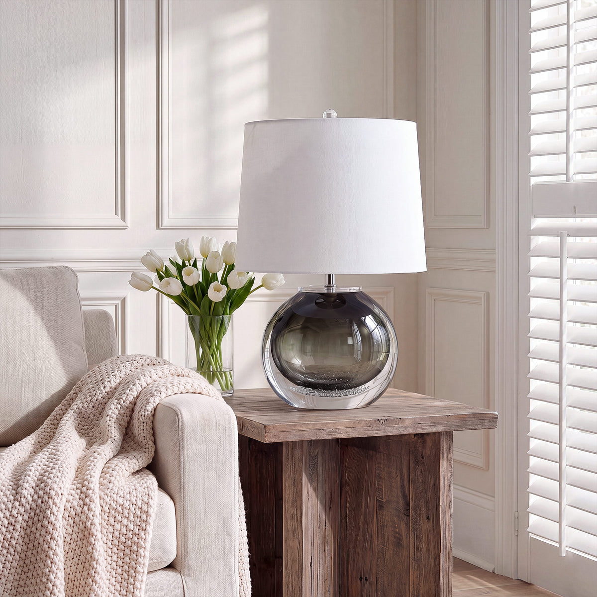 Smoke gray art glass table lamp on a rustic wooden end table next to a sofa with soft blanket