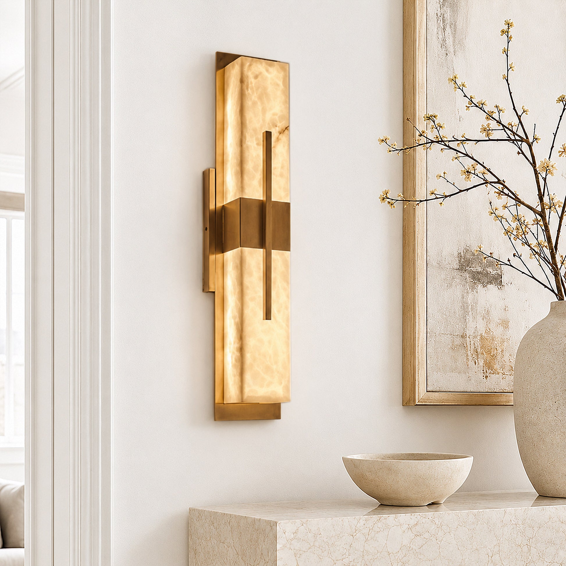 A bright modern entryway showcasing a rectangular brass and alabaster wall sconce mounted on a white textured wall over a travertine console table decorated with a stone bowl and tree branches.