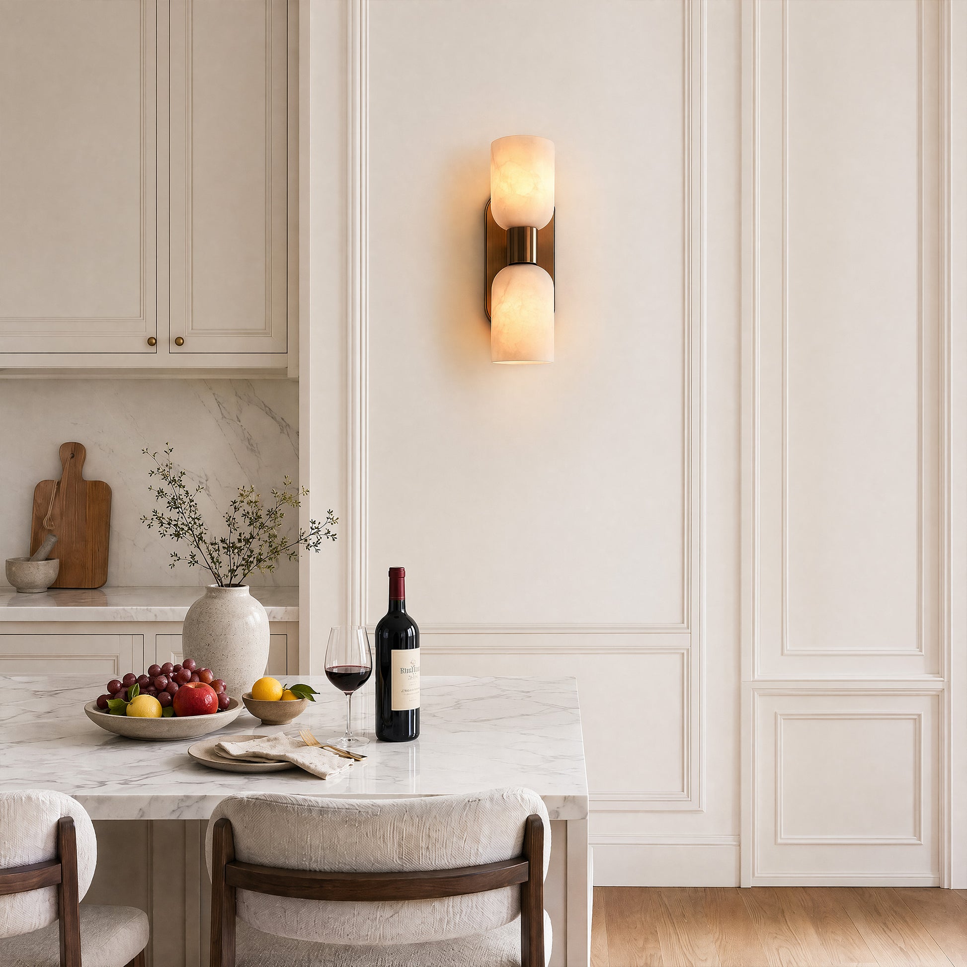 An airy, sunlit kitchen dining space with a white marble island table, wine and fruit platter, and a luxury dual-light alabaster stone wall lamp on a classic wainscoted wall.