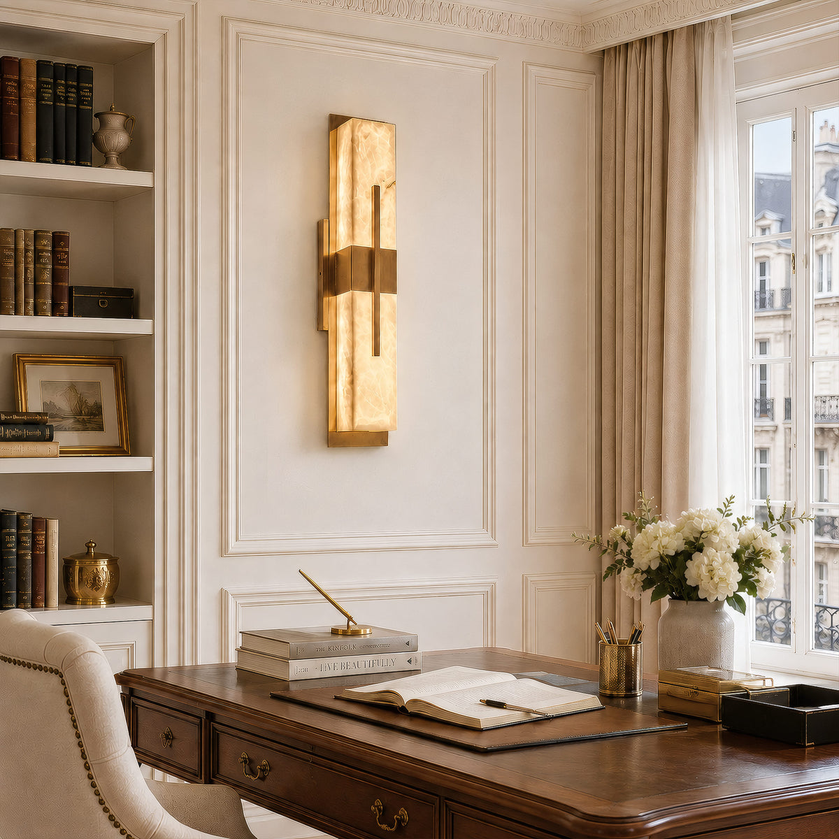 A bright, sunlit Parisian-style home office with a classic wooden writing desk, moldings, white built-in bookshelves, and a vertical geometric alabaster wall sconce by a French window.