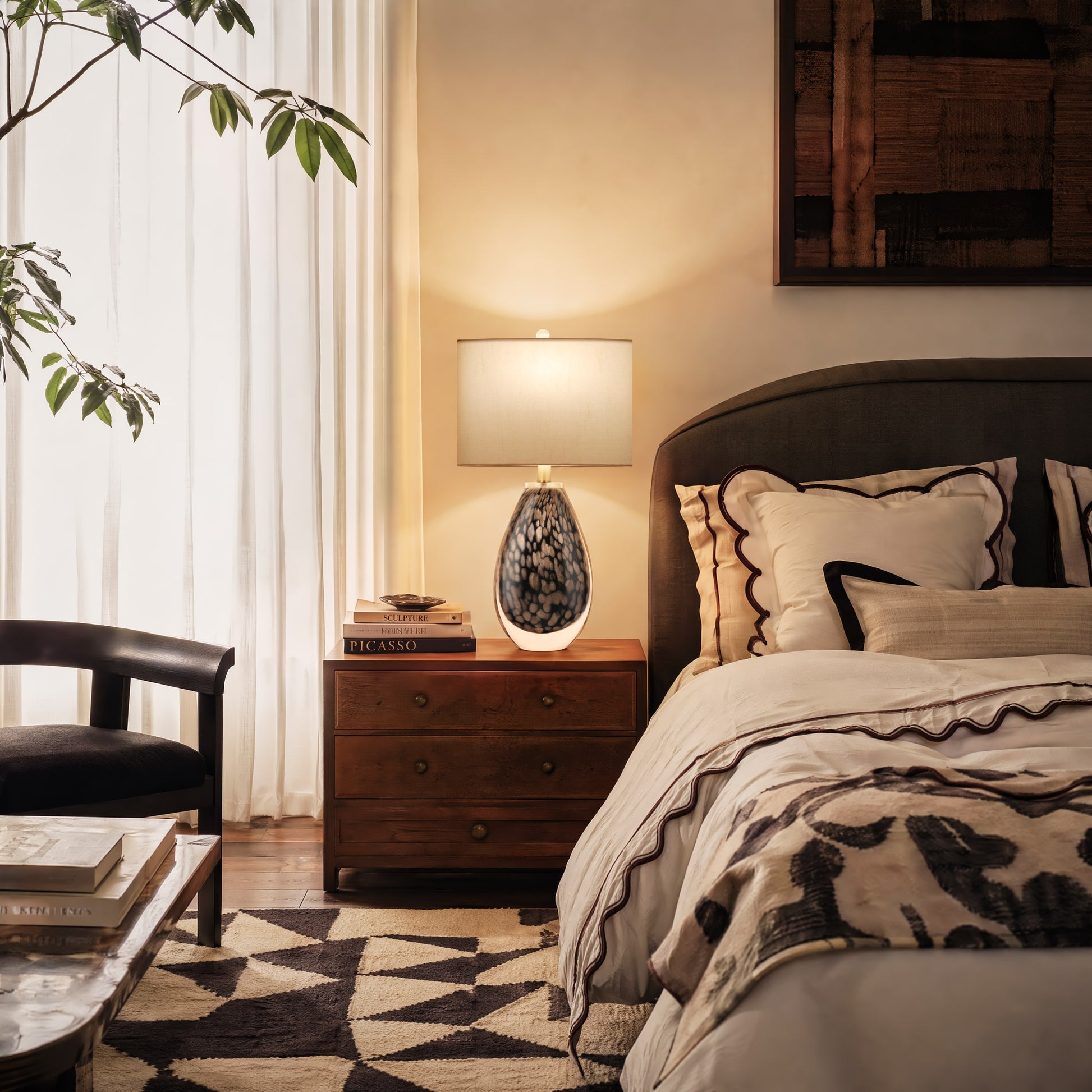 A bright california casual style bedroom featuring a glowing amber scaled glass lamp on a rich walnut nightstand next to geometric pattern rugs and linen bedding.