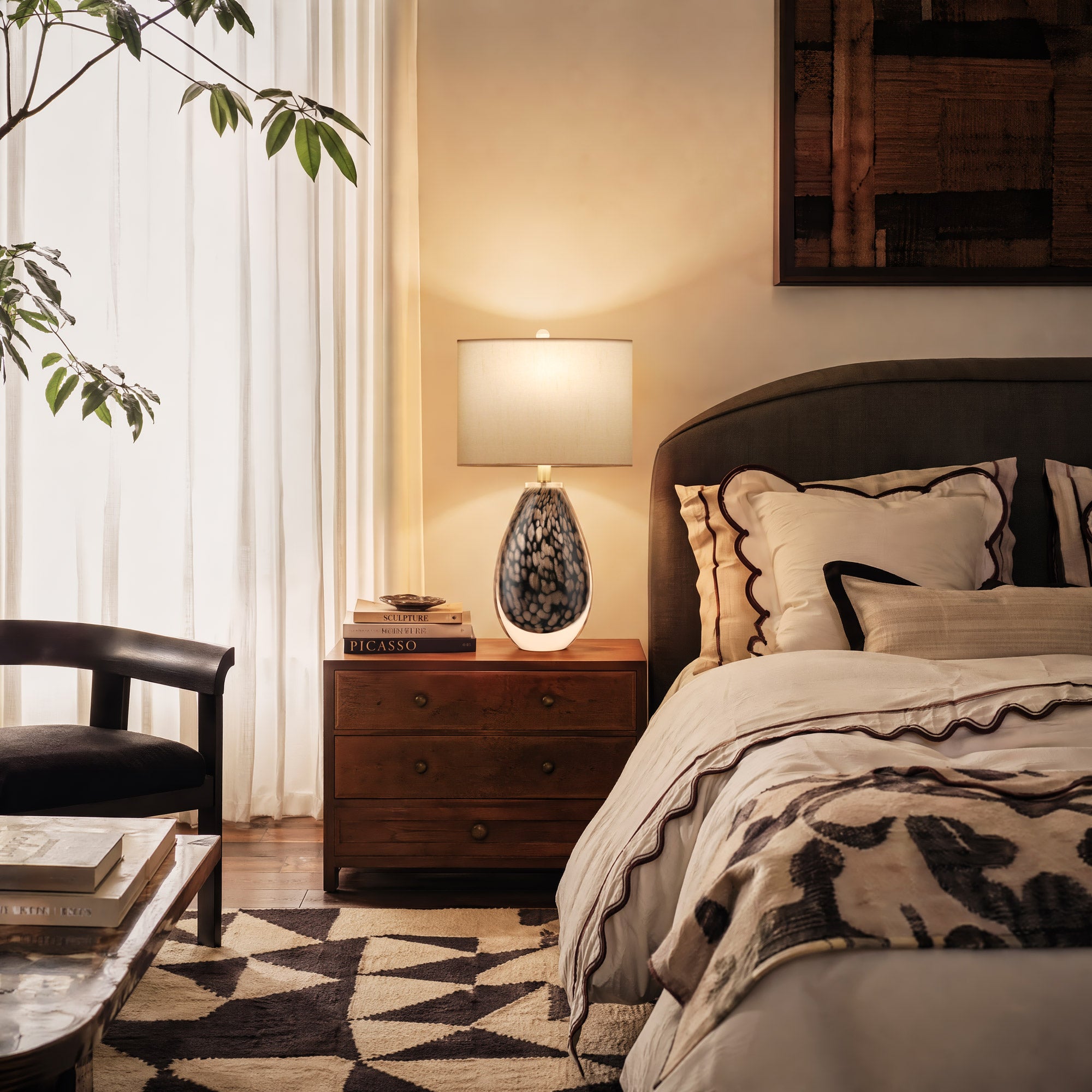 A bright california casual style bedroom featuring a glowing amber scaled glass lamp on a rich walnut nightstand next to geometric pattern rugs and linen bedding.