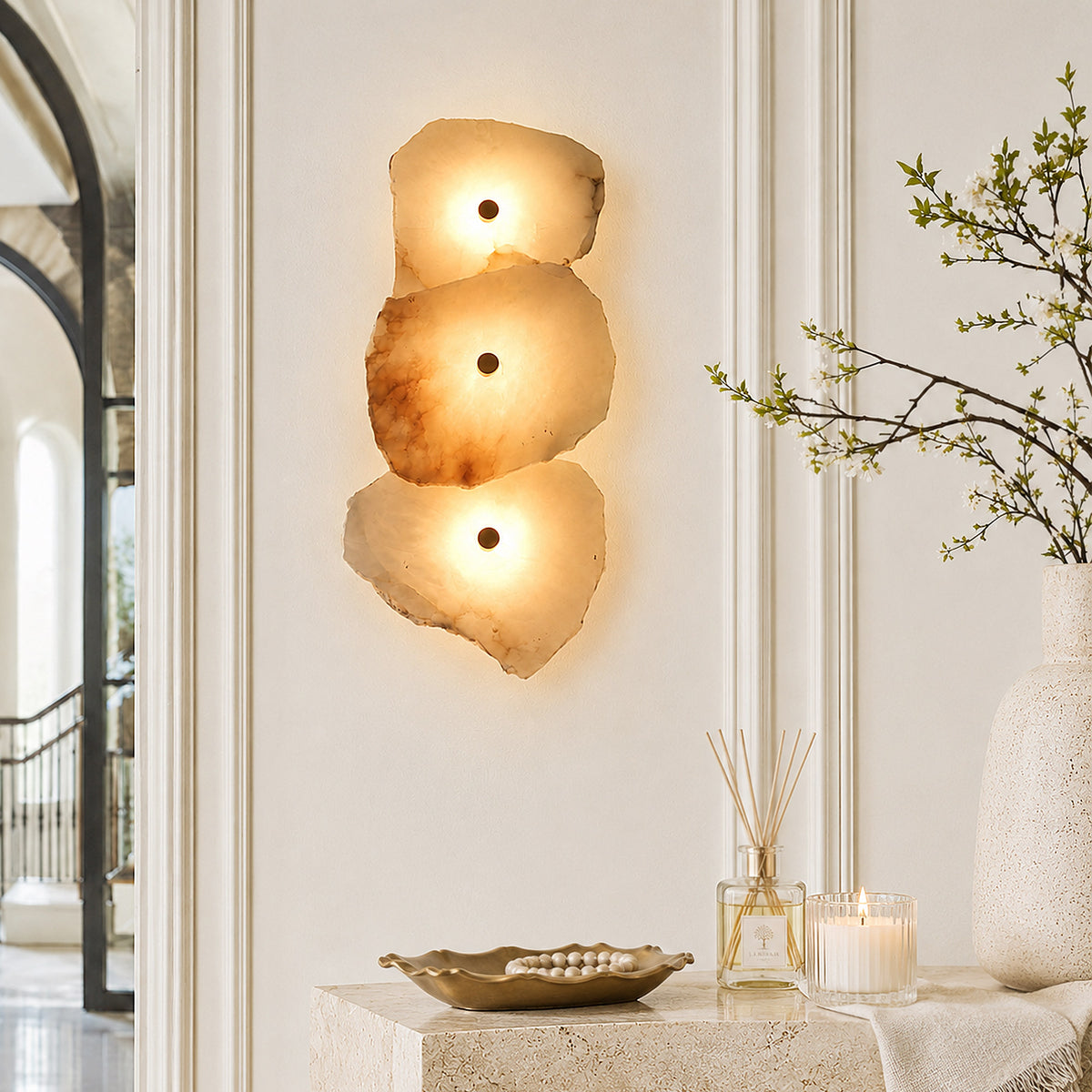 A bright entryway scene featuring a translucent alabaster wall sconce lighting up a travertine console table decorated with a white vase and scented candle.