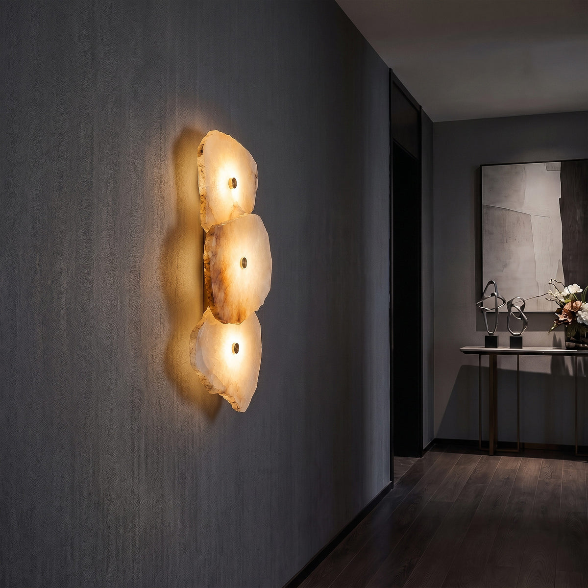 A cinematic, moody modern hallway featuring a 3-light Spanish alabaster wall sconce emitting a warm amber glow on a textured dark grey plaster wall, leading to an entryway console table with metal sculptures.