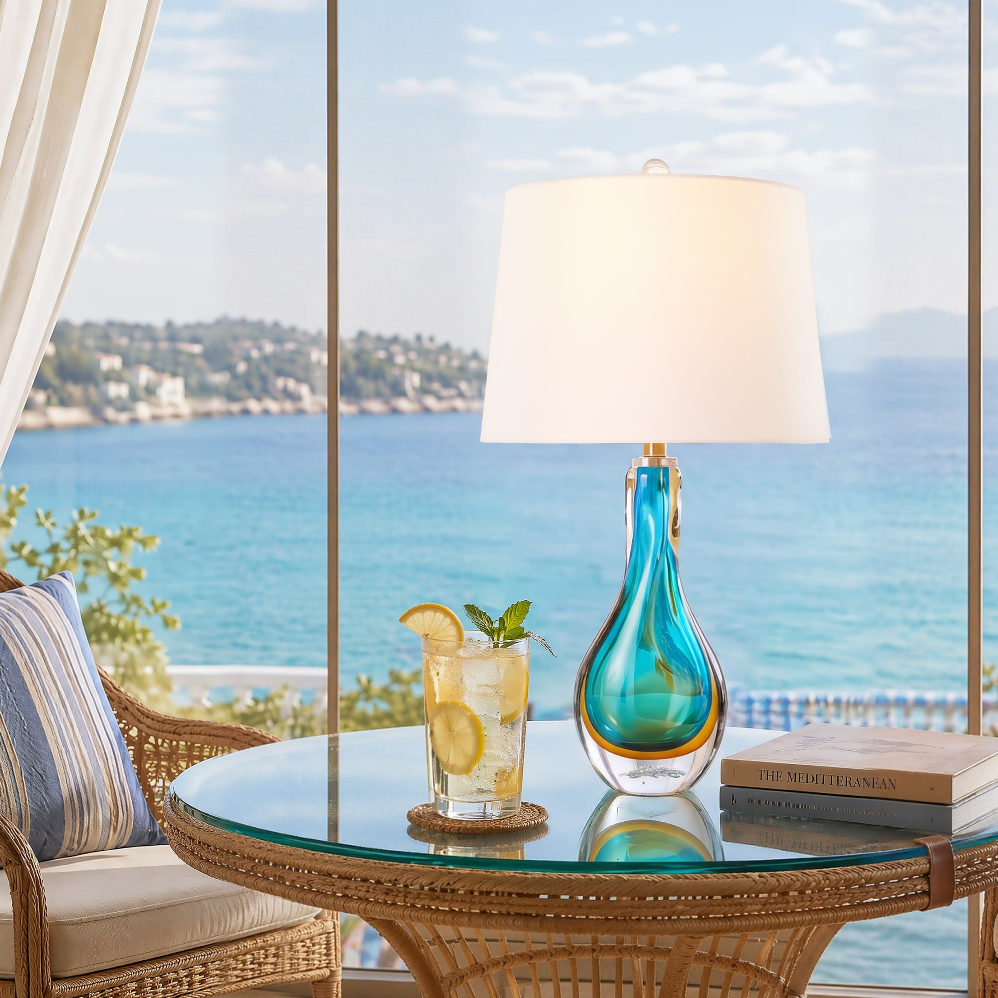 A luxury seaside villa sunroom or patio featuring a lit turquoise blown glass lamp on a rattan glass table next to a refreshing cocktail facing the blue Mediterranean sea view.