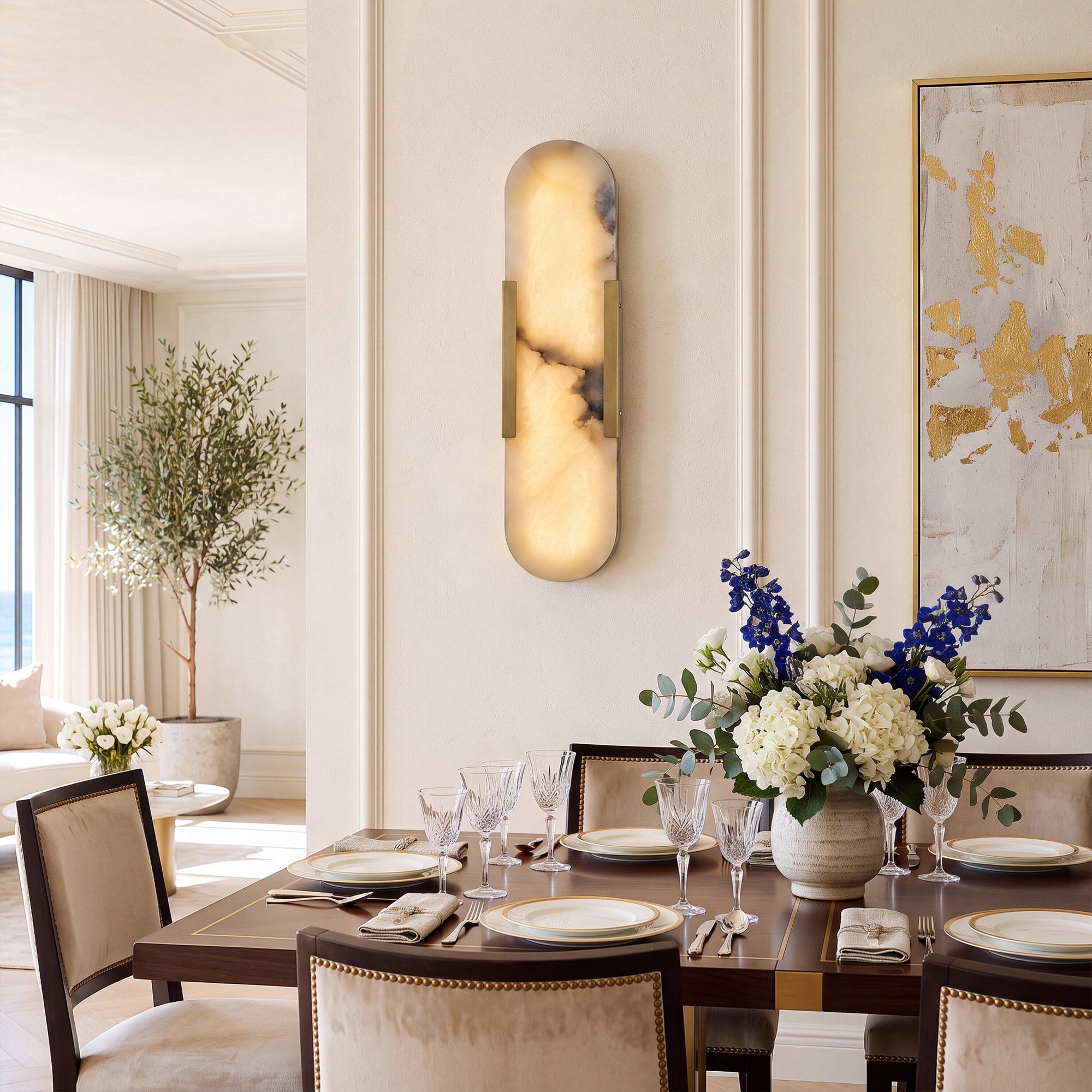 An elegant, creamy tone dining room highlighting a capsule alabaster wall lamp on a paneled wall, above a formal dark wood dining table beautifully set with blue and white flowers.