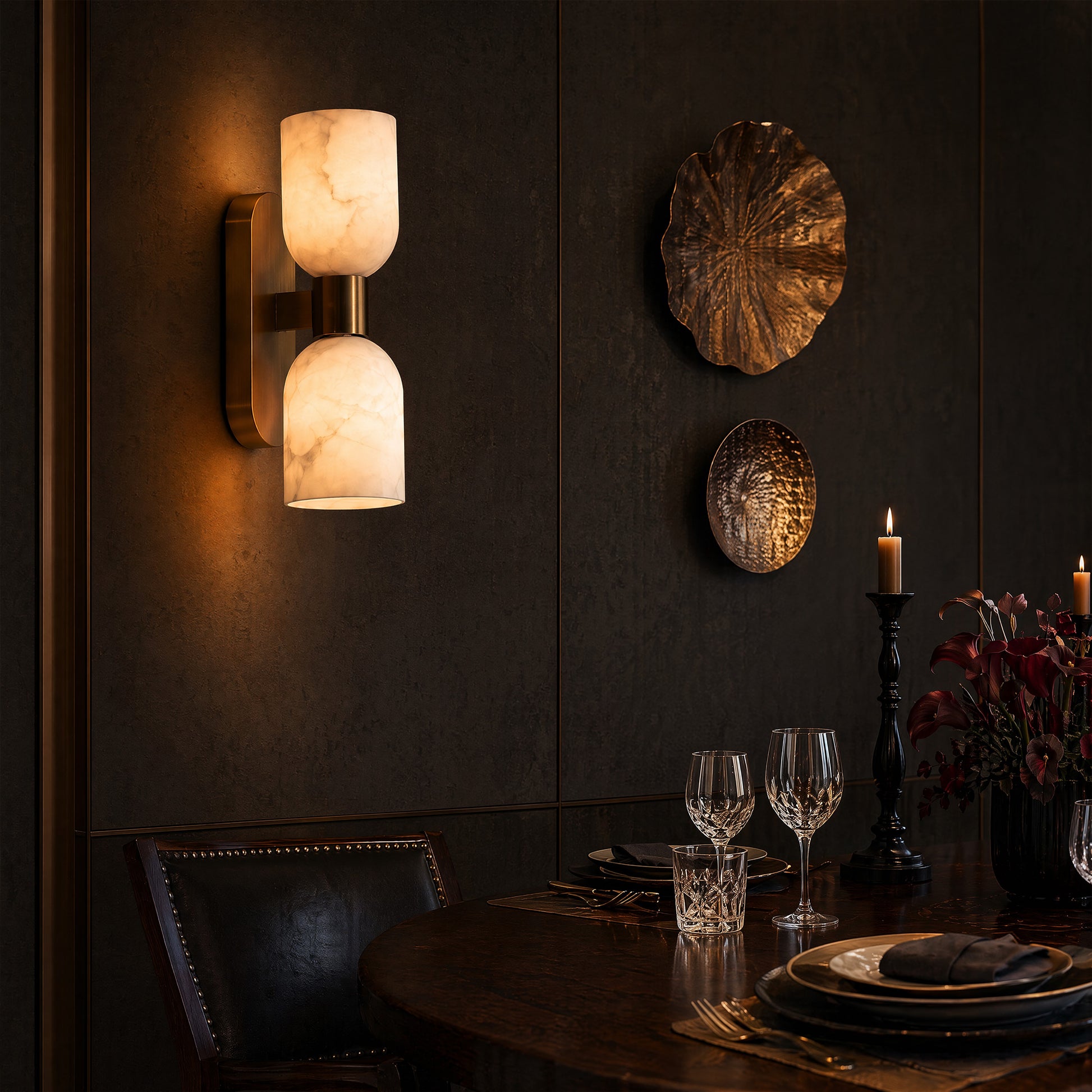 A moody, high-end dining room featuring a glowing double-headed alabaster stone wall sconce on a dark dark-brown paneled wall, above a wooden dining table set with wine glasses.