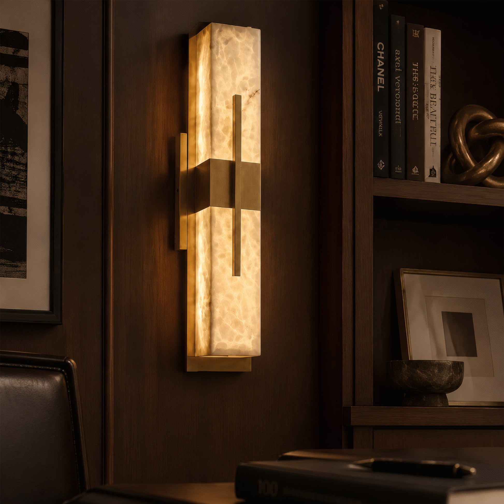 An upscale, moody study room with dark wood paneling, featuring a glowing linear brass and alabaster wall sconce next to a bookshelf curated with luxury art books.