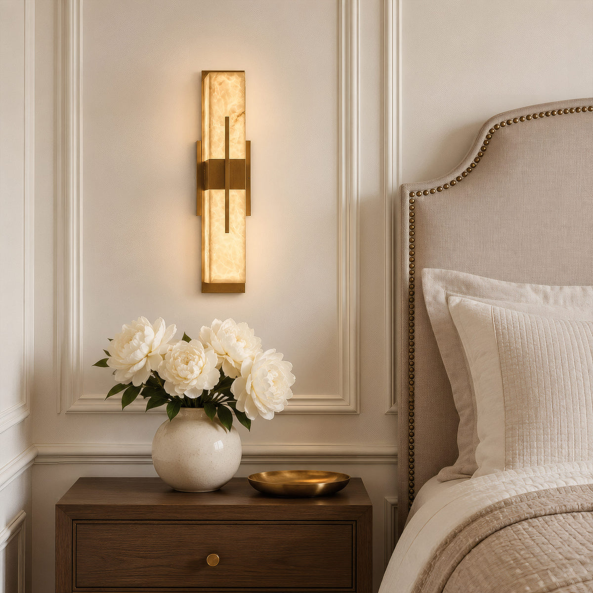 A serene, neutral-toned master bedroom featuring a linear brass alabaster wall light mounted next to a beige tufted headboard and a wooden nightstand with fresh white peonies.